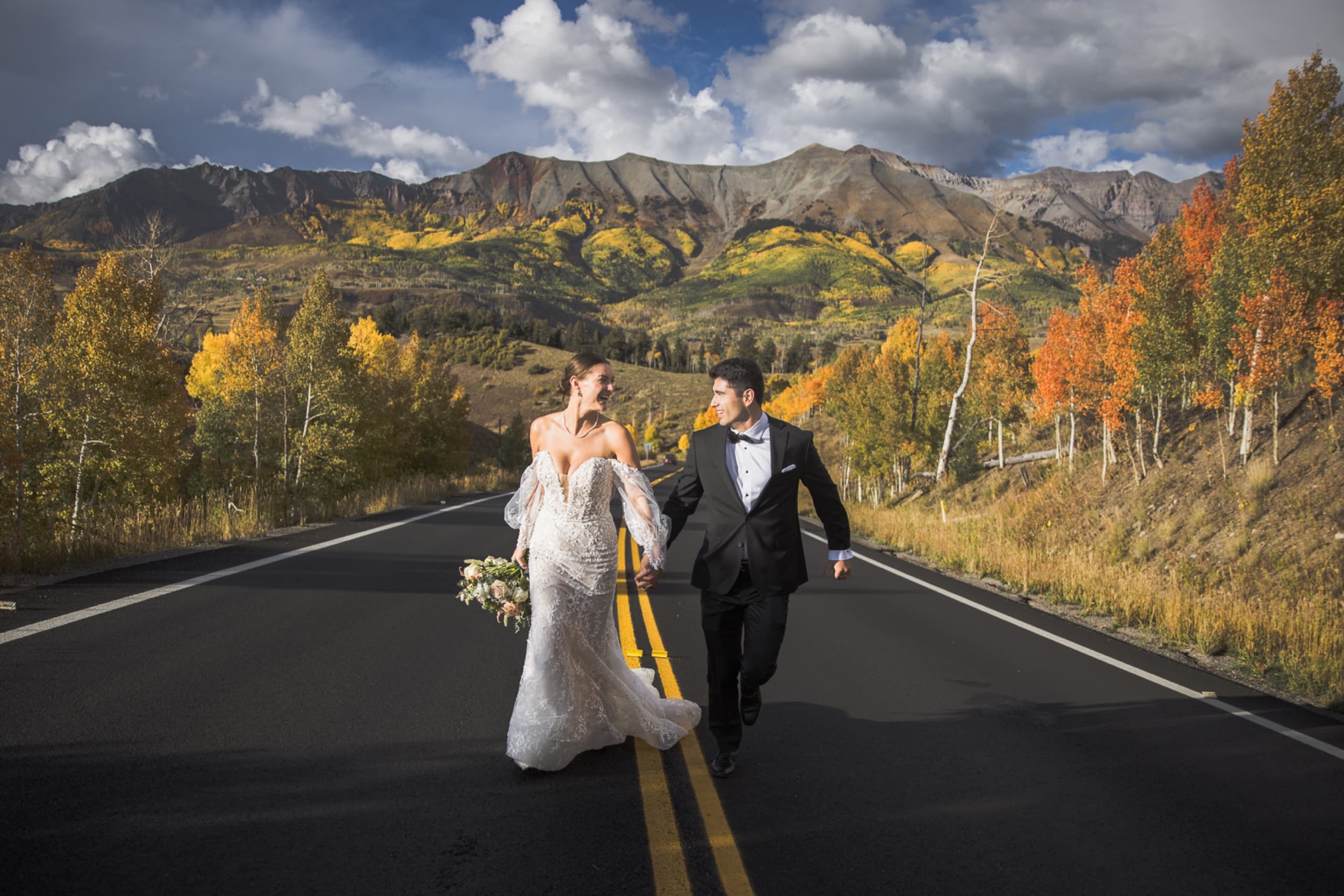 Bride and groom walking hand in hand down a mountain highway in vibrant fall foliage in Telluride, Colorado