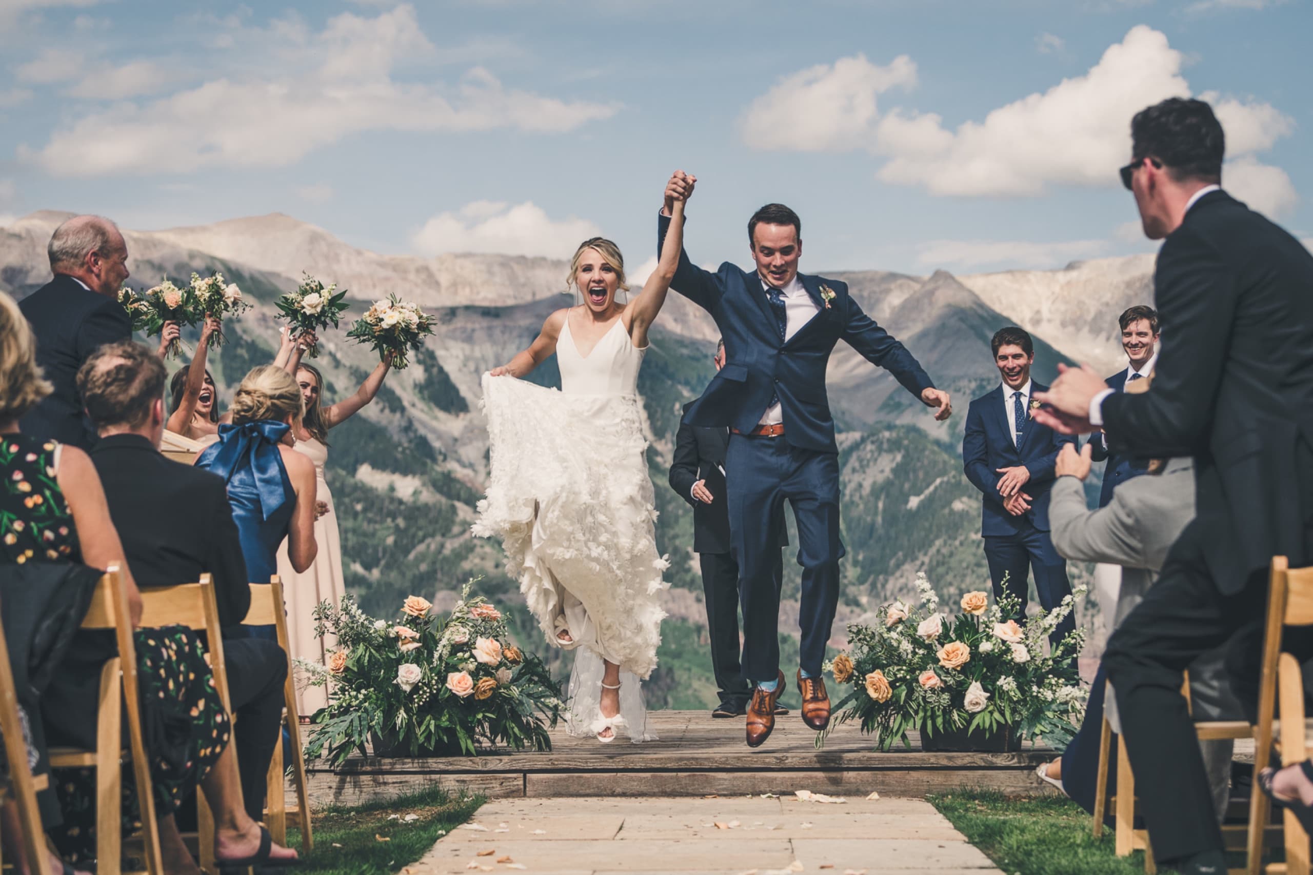 Bride and groom jumping together as they walk the aisle with Telluride mountain peaks behind them — alpine weddings