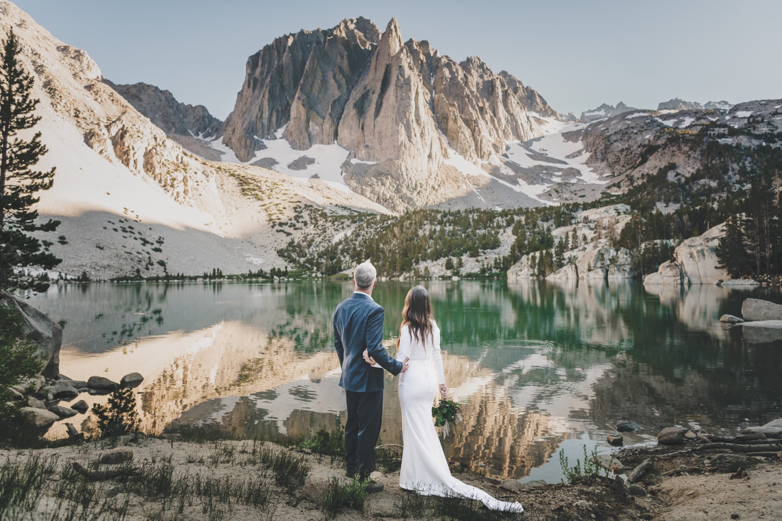 Couple hand in hand before a mirror-still alpine lake reflecting jagged Sierra Nevada peaks