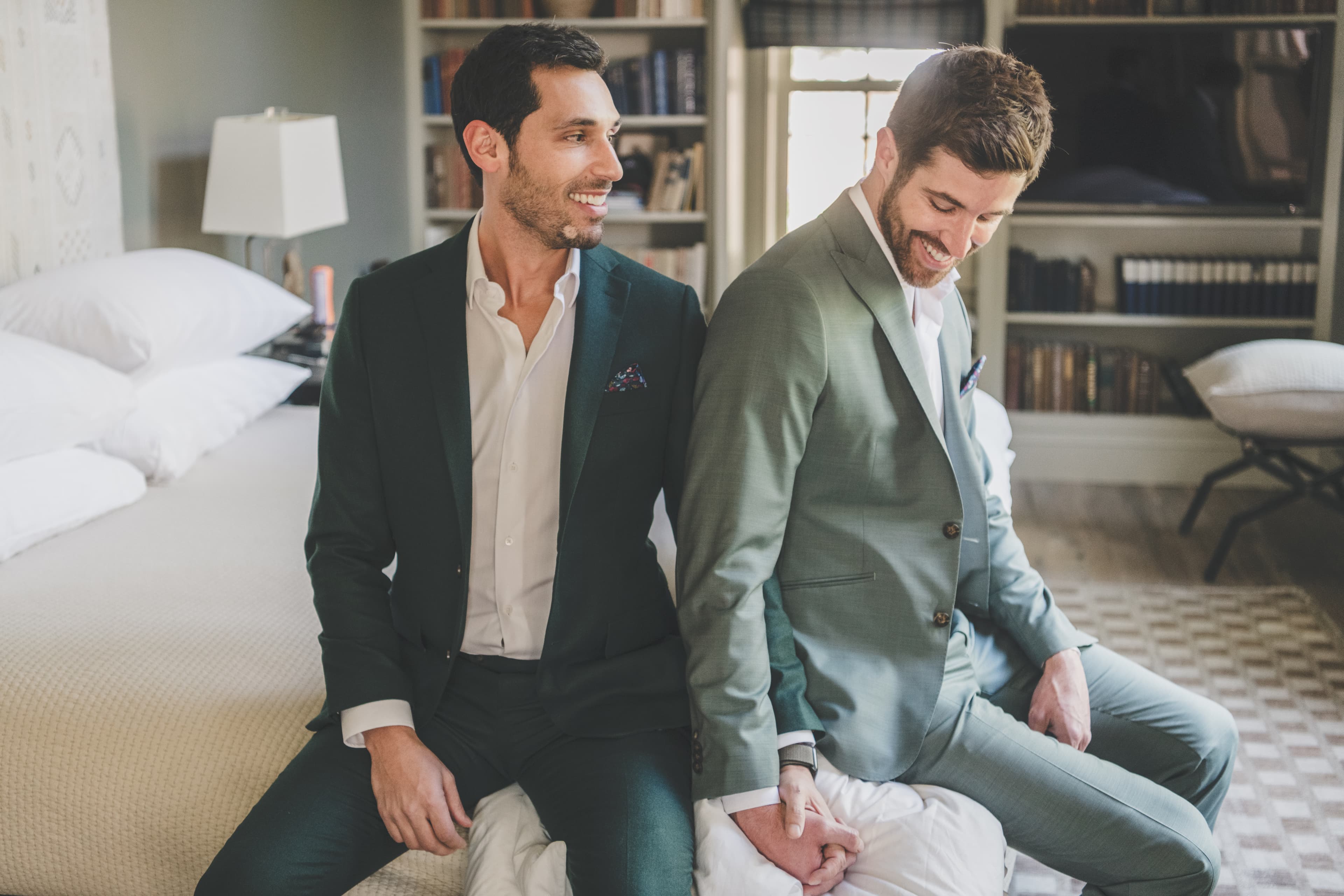 Greg and John sit back-to-back in their bridal suite, holding hands behind them as they prepare for their wedding