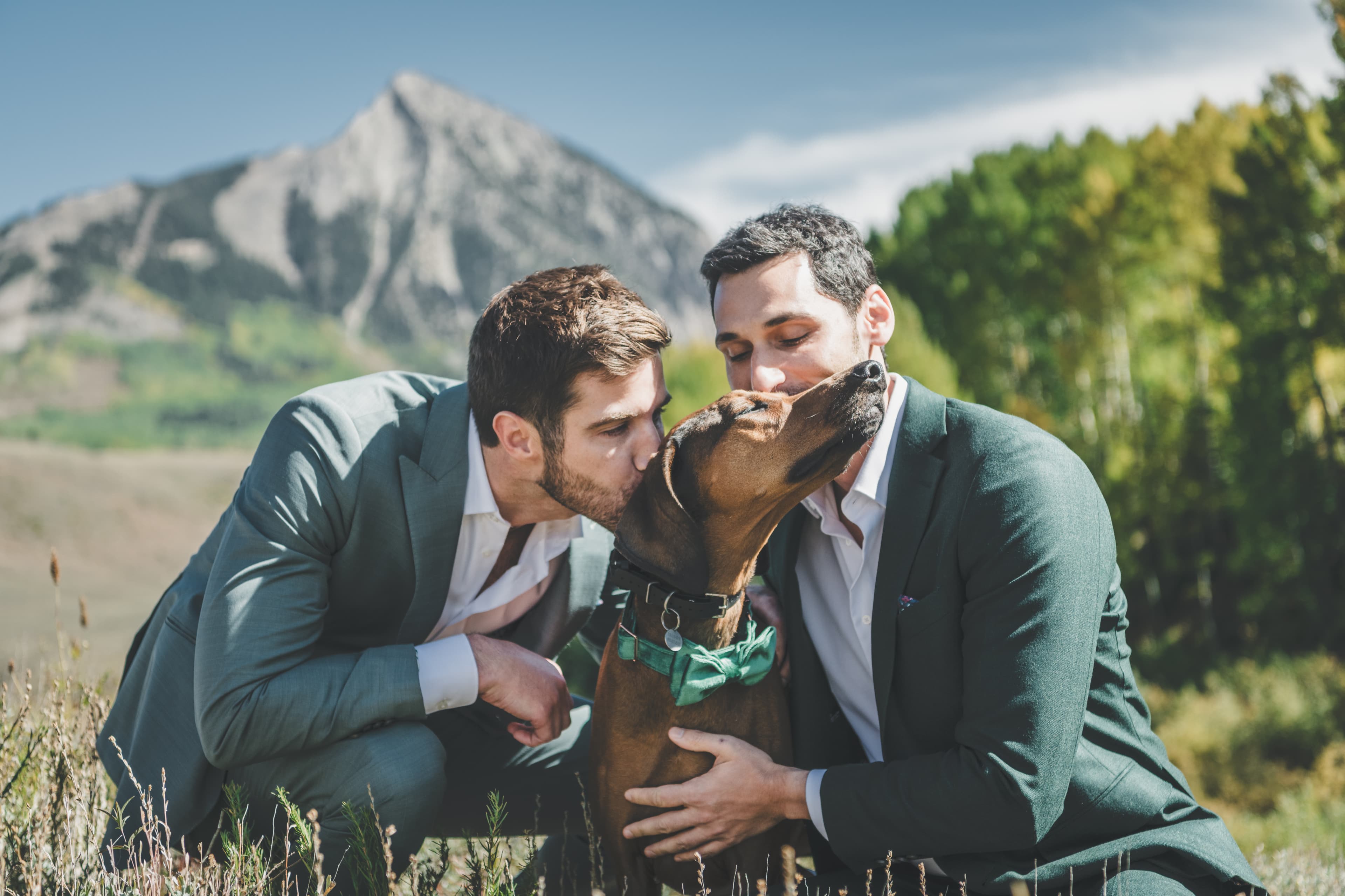 Greg and John share a kiss with their dog wearing a green bowtie, Mt. Crested Butte rising behind them