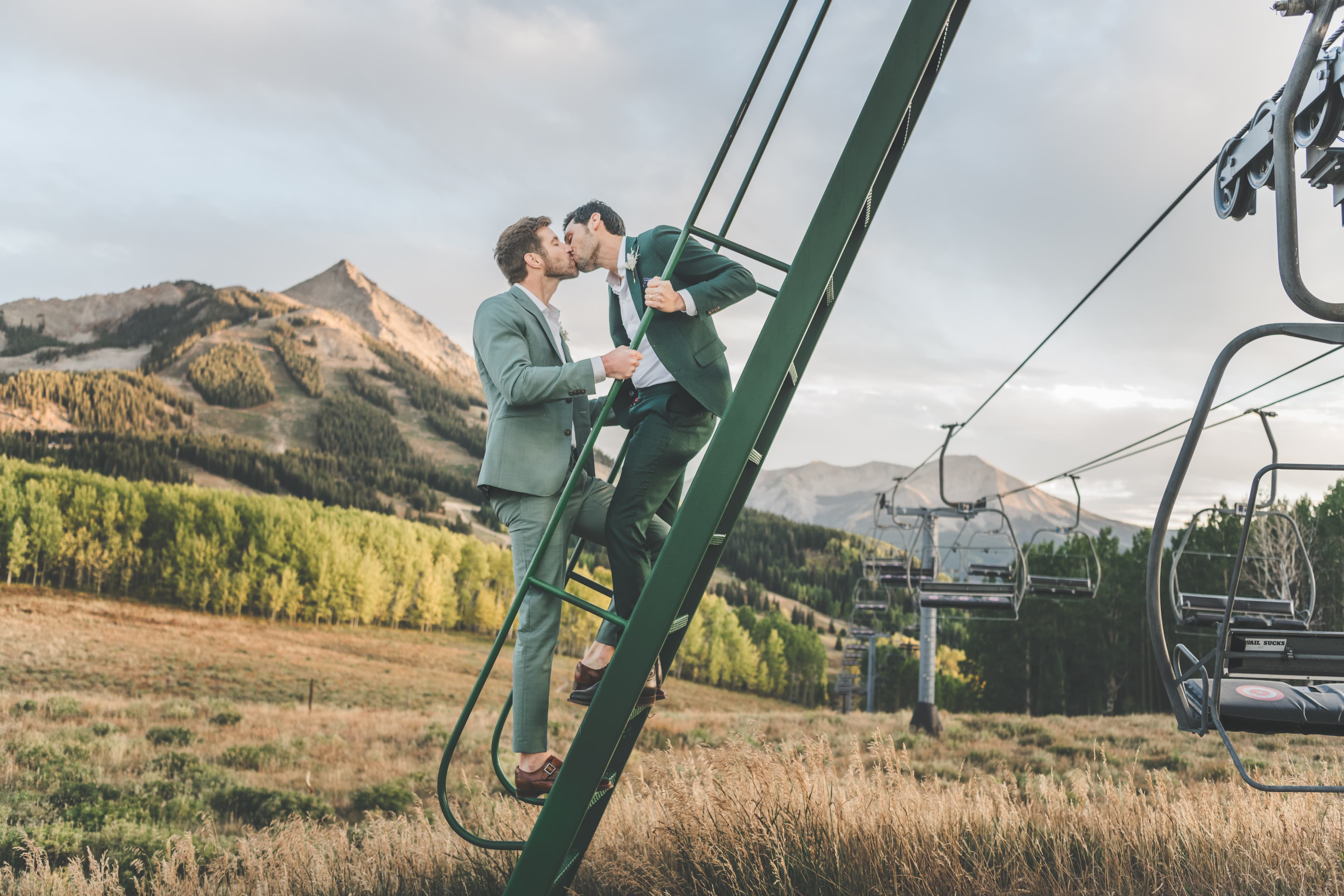 Greg and John kiss on a chairlift tower with Mt. Crested Butte rising behind them at their Ten Peaks Lodge wedding in Crested Butte, Colorado