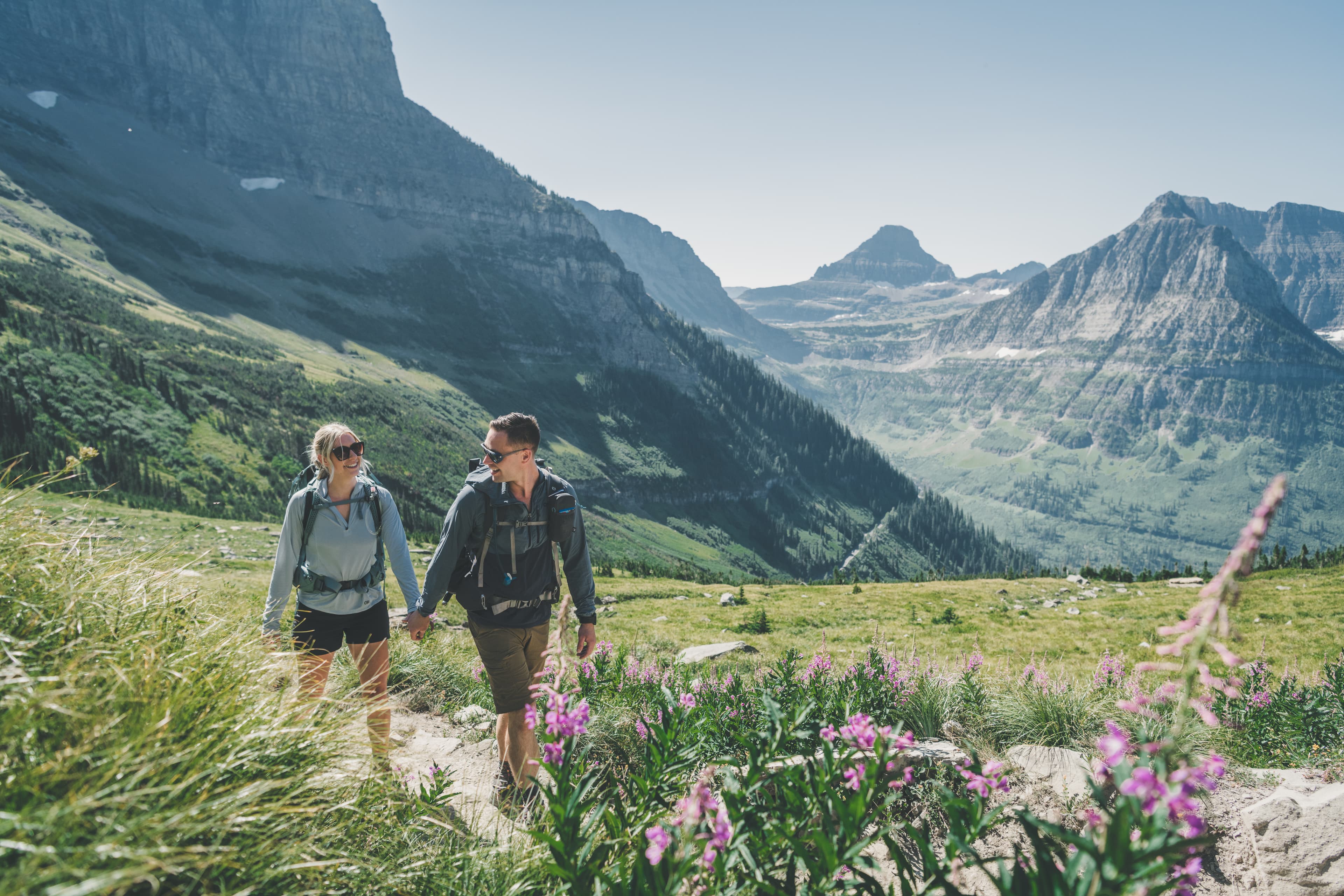 Shawna and Ryan hiking hand-in-hand through fireweed and Glacier National Park alpine meadows