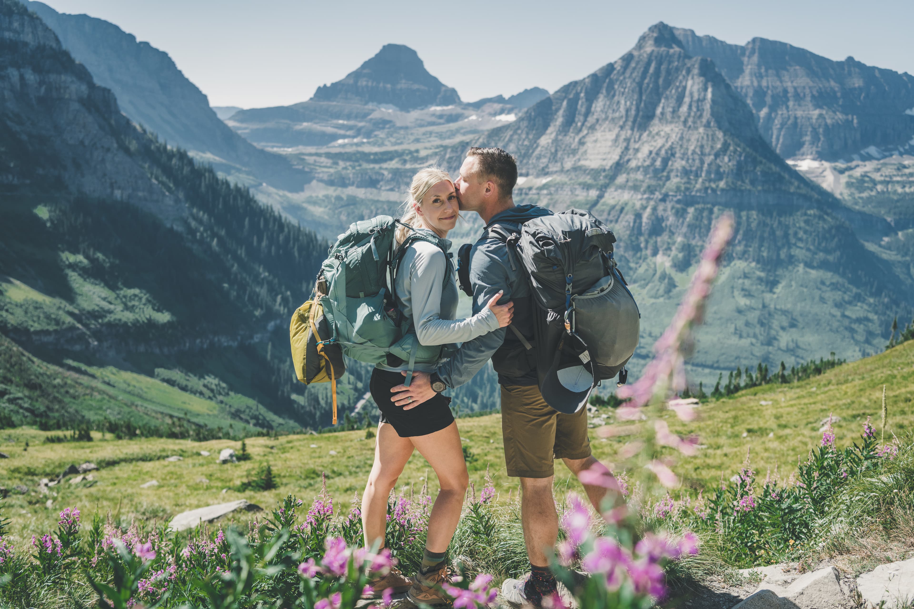 Shawna and Ryan kissing in their packs above Logan Pass with fireweed and Glacier peaks behind them