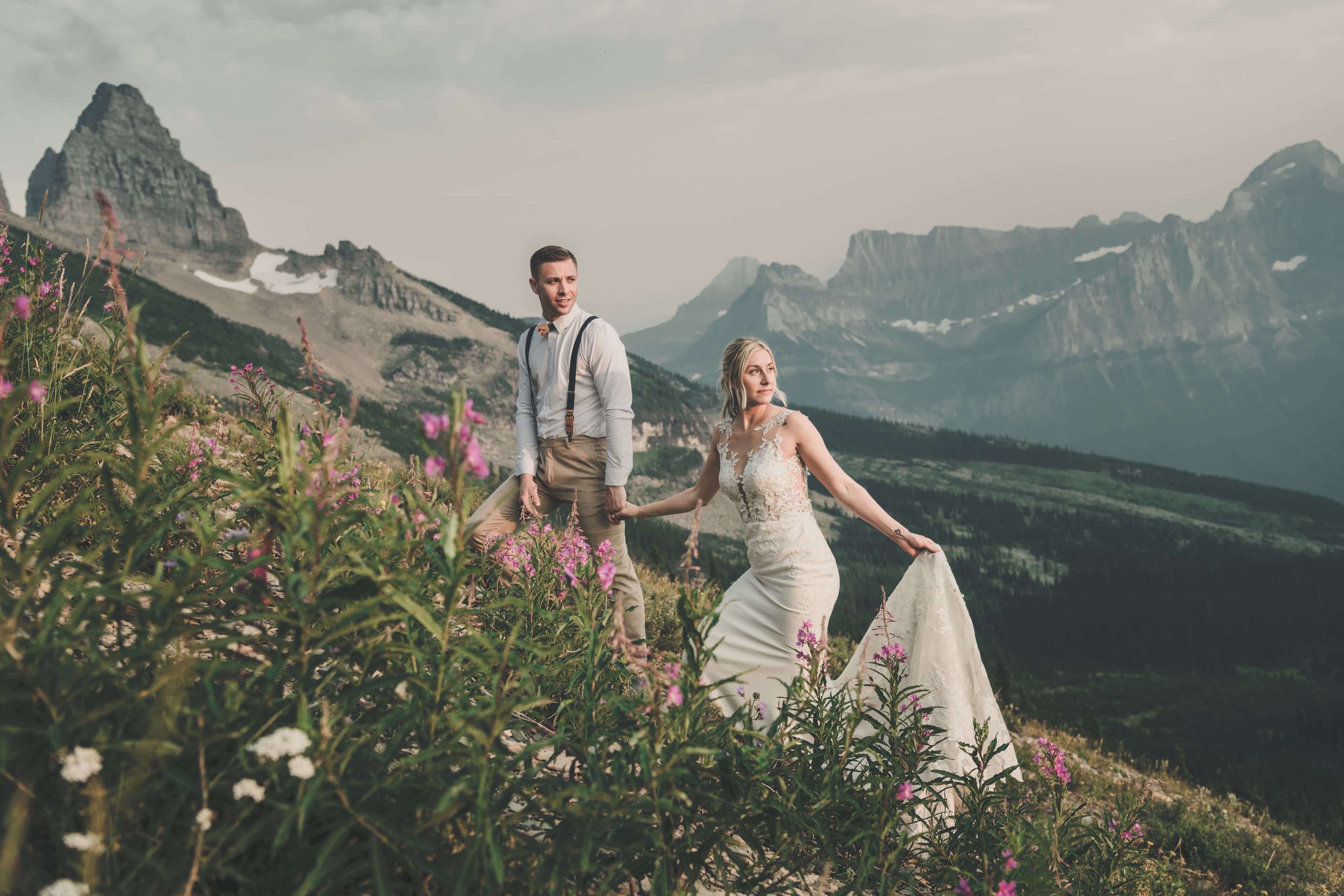Shawna walking through fireweed in her gown with Ryan close behind, Glacier peaks dramatic behind them