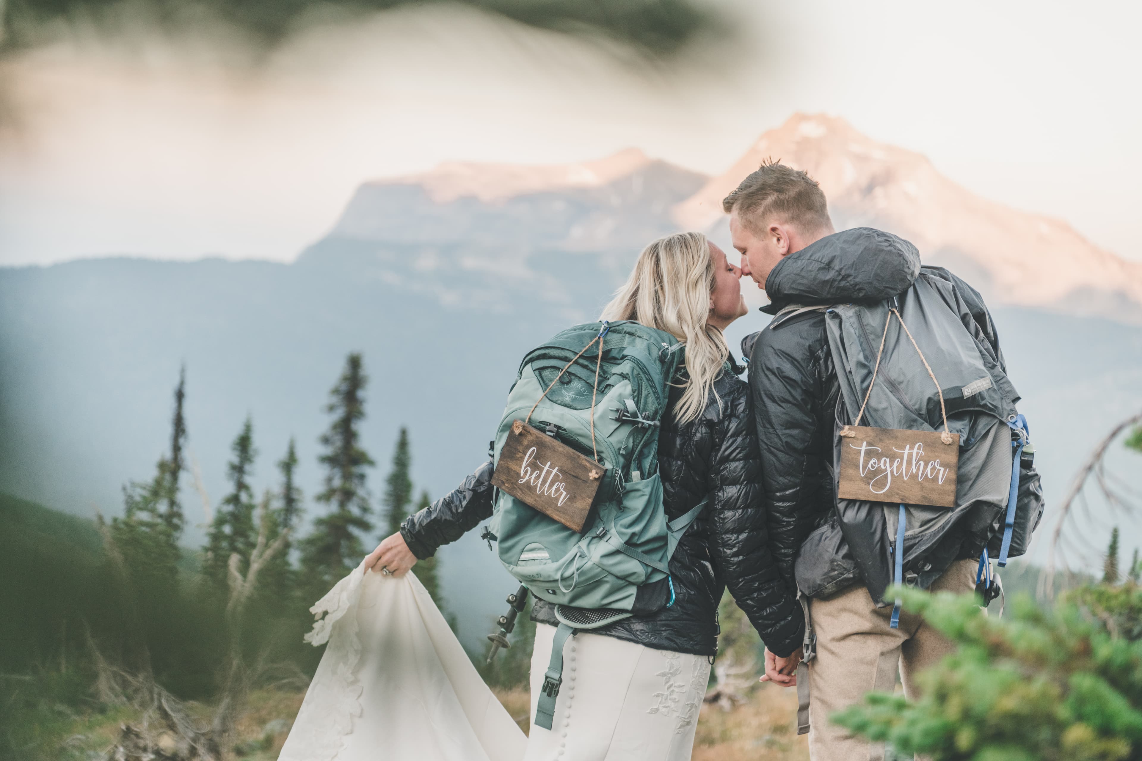Shawna and Ryan kissing in front of an alpenglow Glacier peak with their 'Better' 'Together' wood-sign backpacks