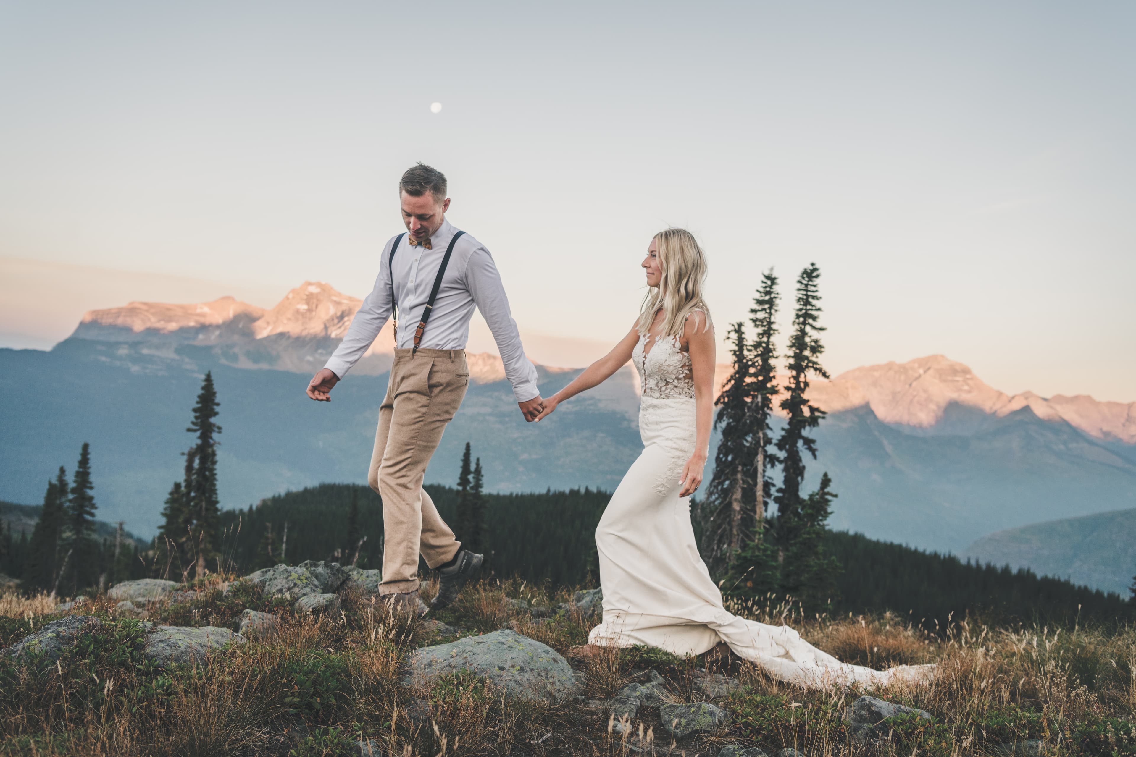 Shawna and Ryan walking through dusk meadow with the moon rising over the Glacier ridgeline behind them
