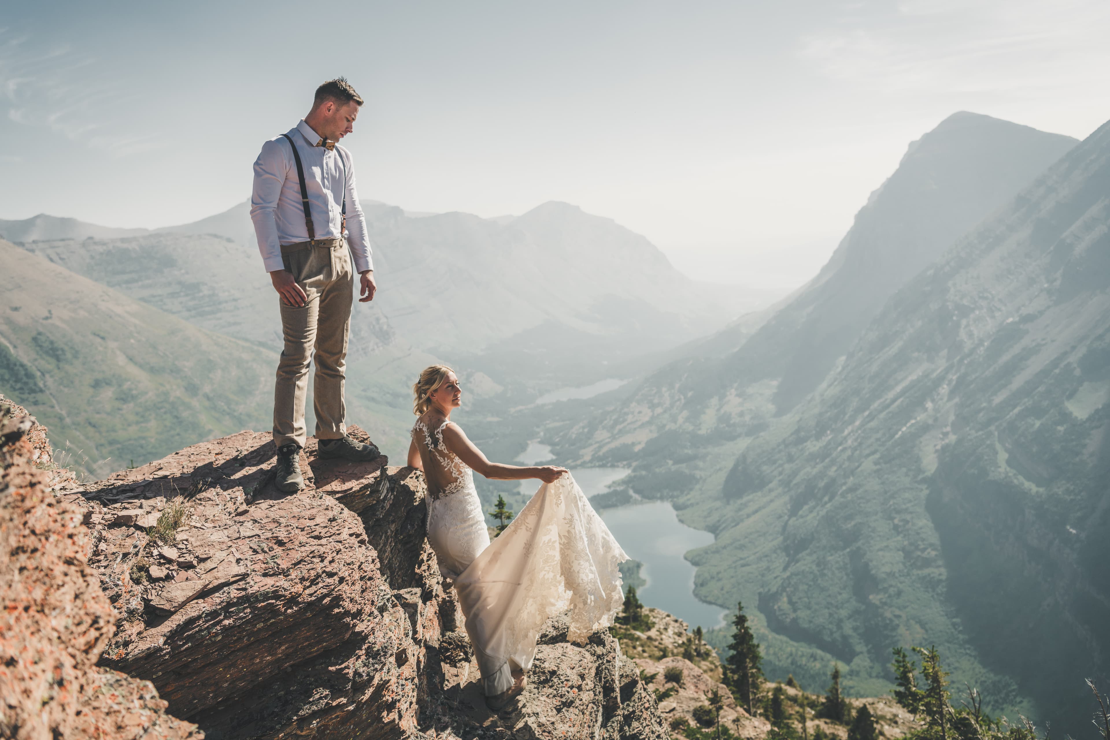 Ryan watching Shawna kneel at the cliff edge with her gown trailing toward the lake-filled Many Glacier valley below