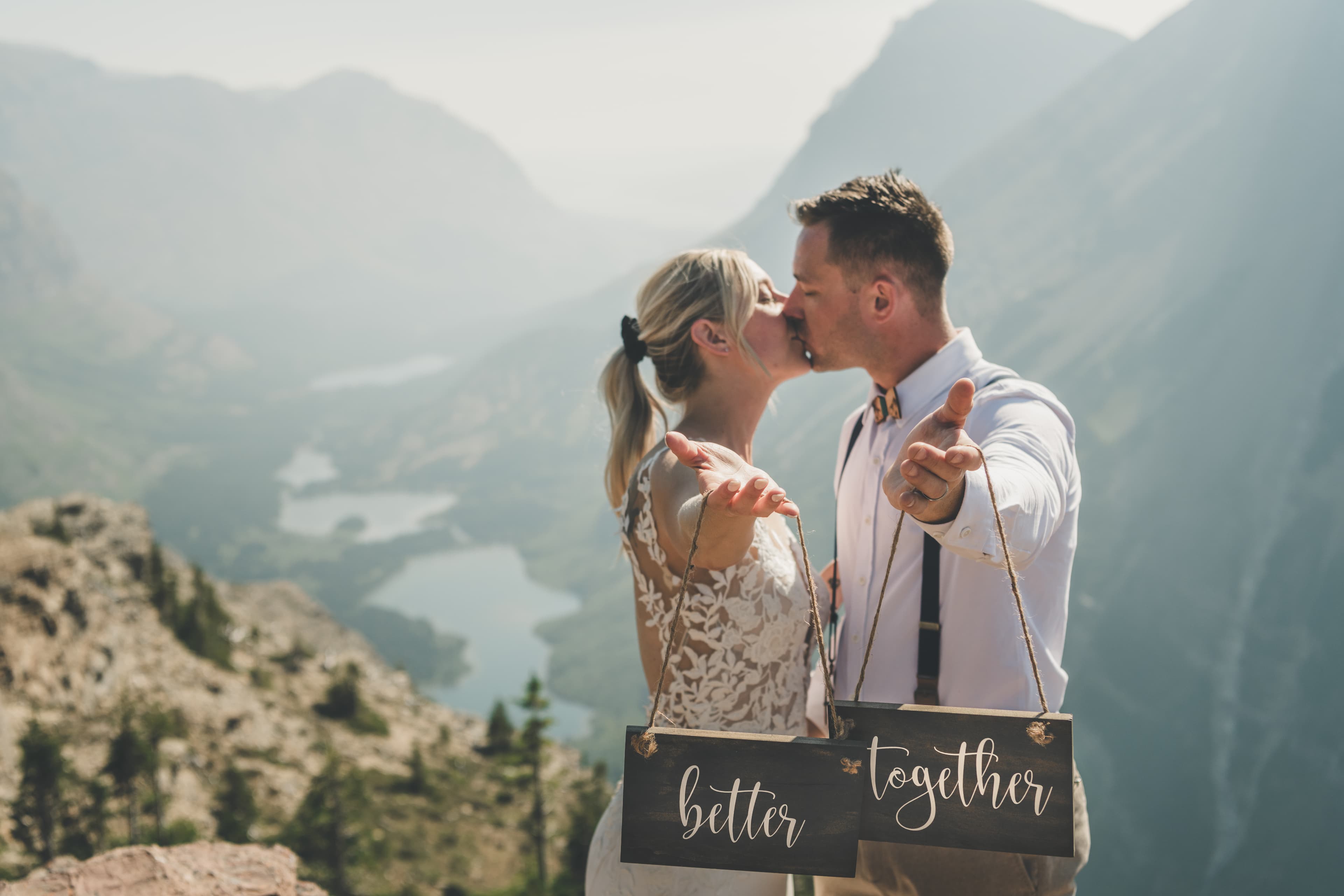 Shawna and Ryan kissing while holding 'Better' and 'Together' signs at a cliff above the Many Glacier valley lakes