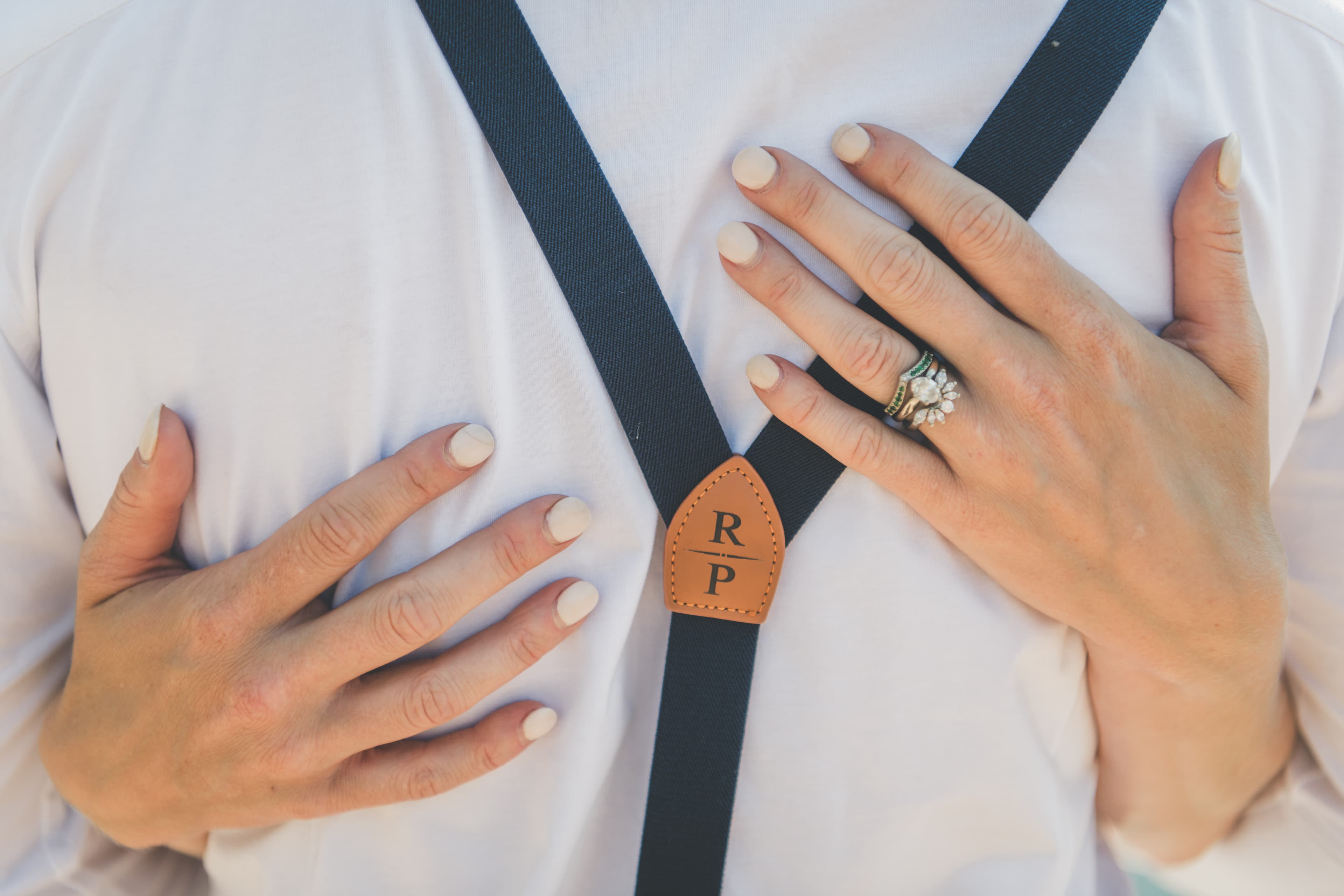 Detail close-up of Shawna's wedding rings and ivory manicure resting on Ryan's chest beside his monogrammed R/P suspender clip