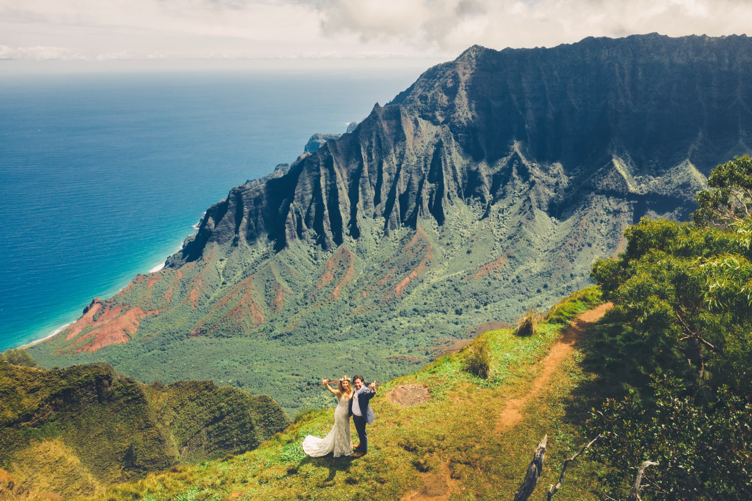 Aerial view of Erin and Kenny standing tiny against the Na Pali Coast cliffs from the Kalalau Lookout