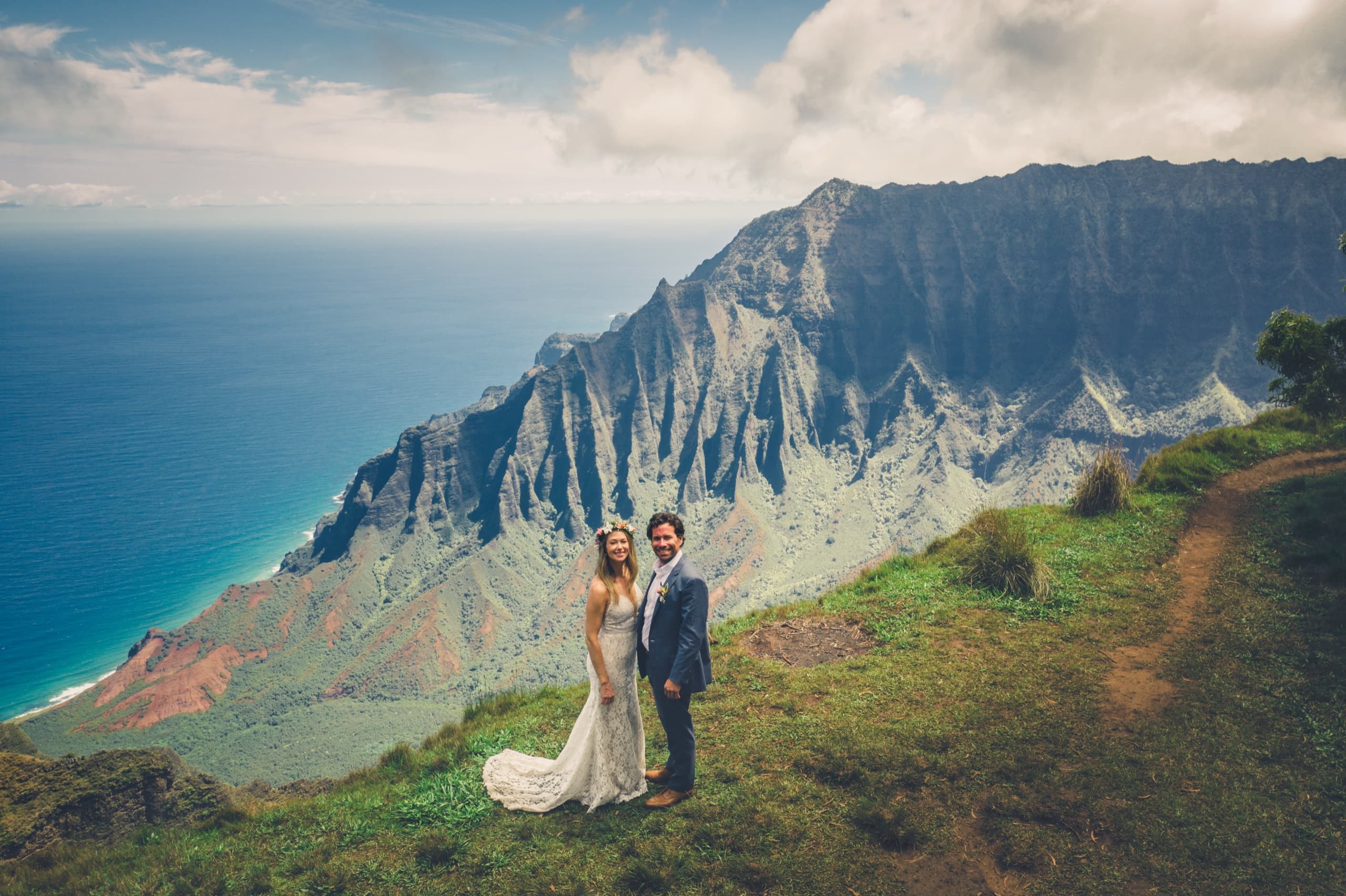 Drone shot of Erin and Kenny on the grassy edge of the Kalalau Lookout with Na Pali laid out beneath them