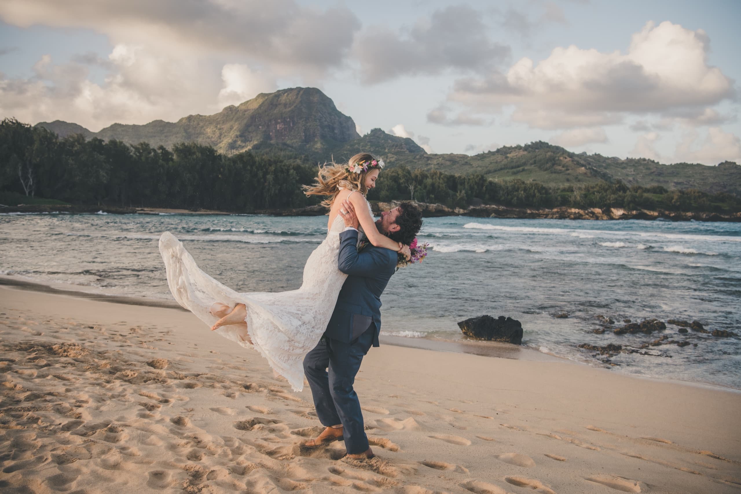 Kenny lifts Erin off her feet on the beach at golden hour with Hāʻupu Ridge across the bay