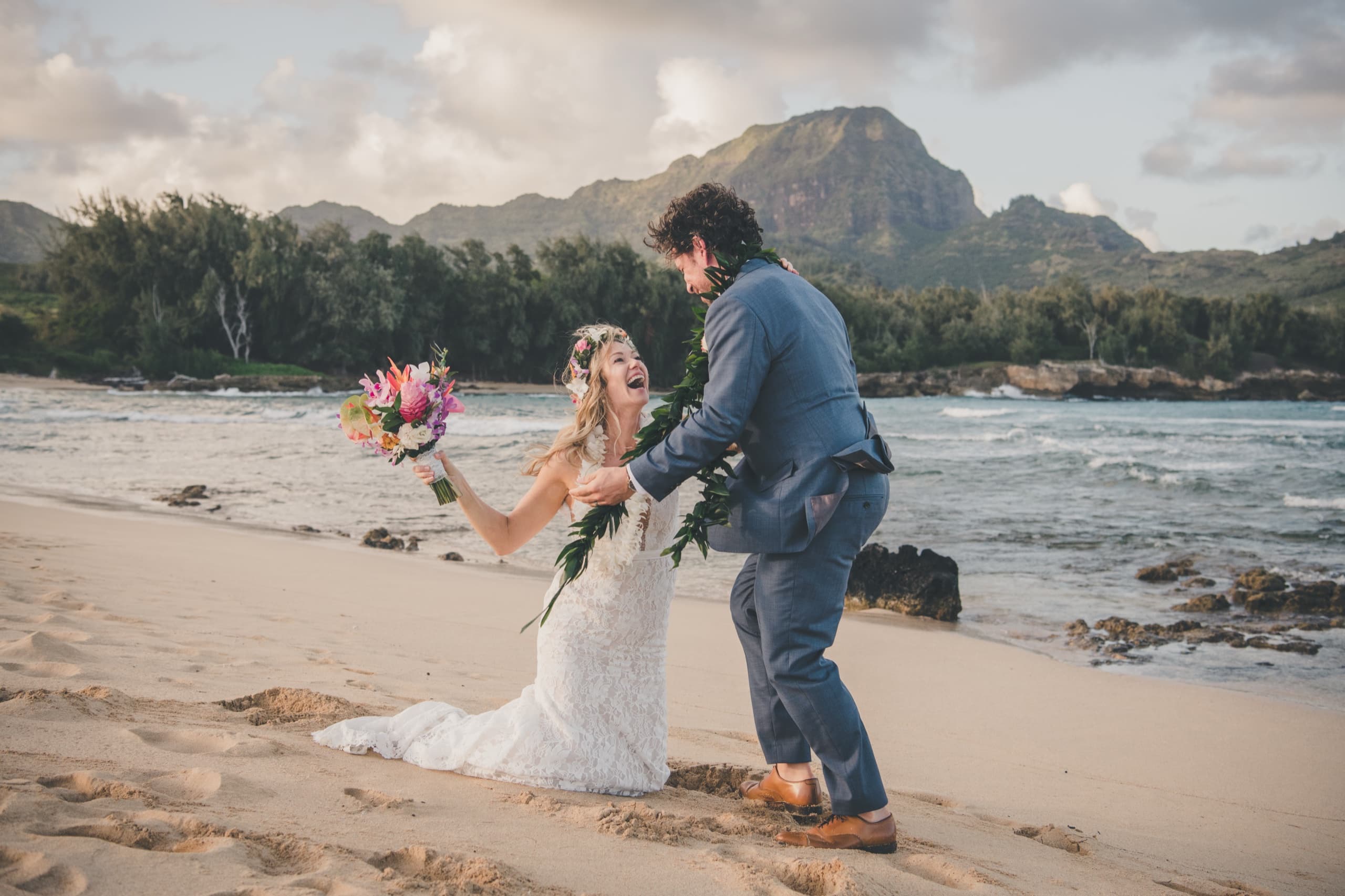 Erin laughing mid-step on the beach with her bouquet, mountains and surf behind her