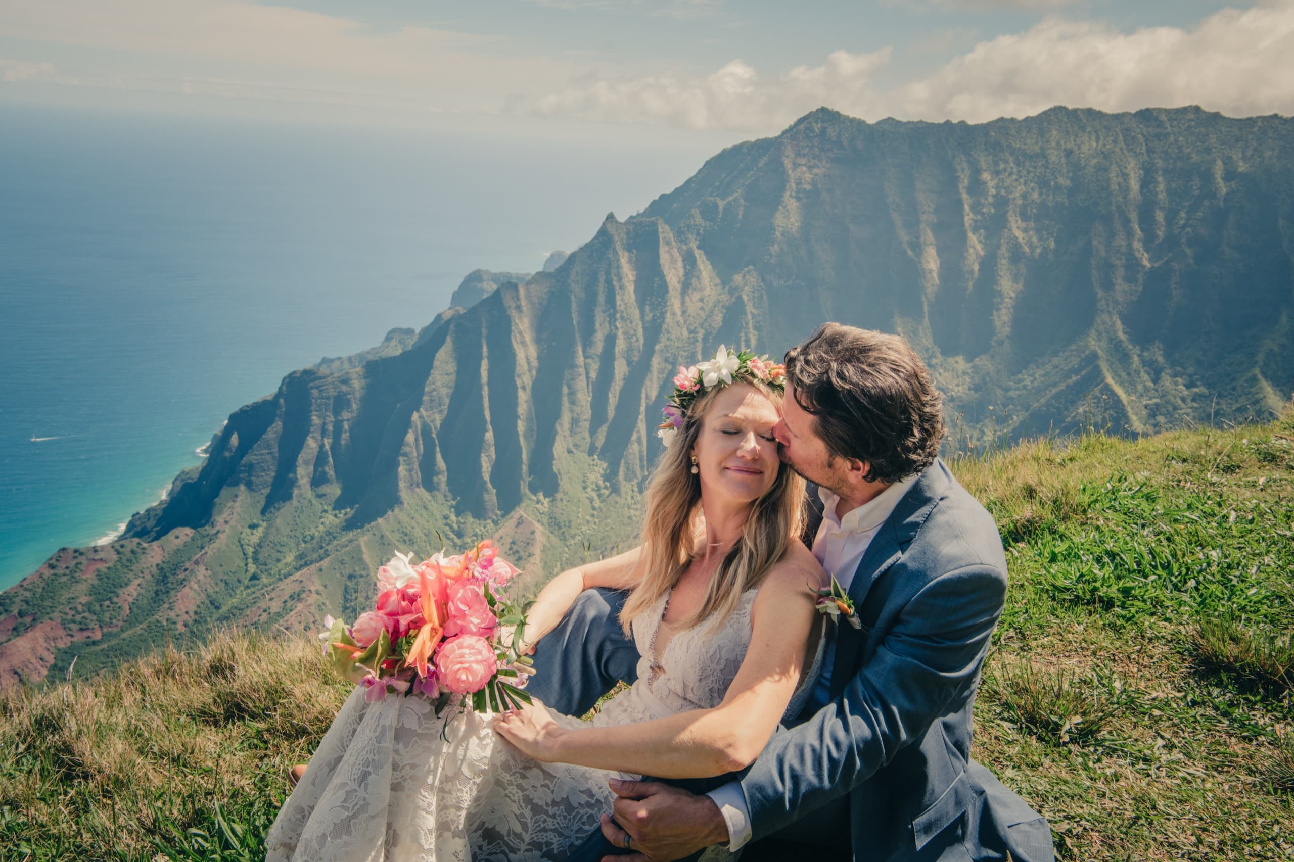 Erin and Kenny seated on the grass at the Kalalau Lookout, Kenny kissing her forehead with Na Pali below