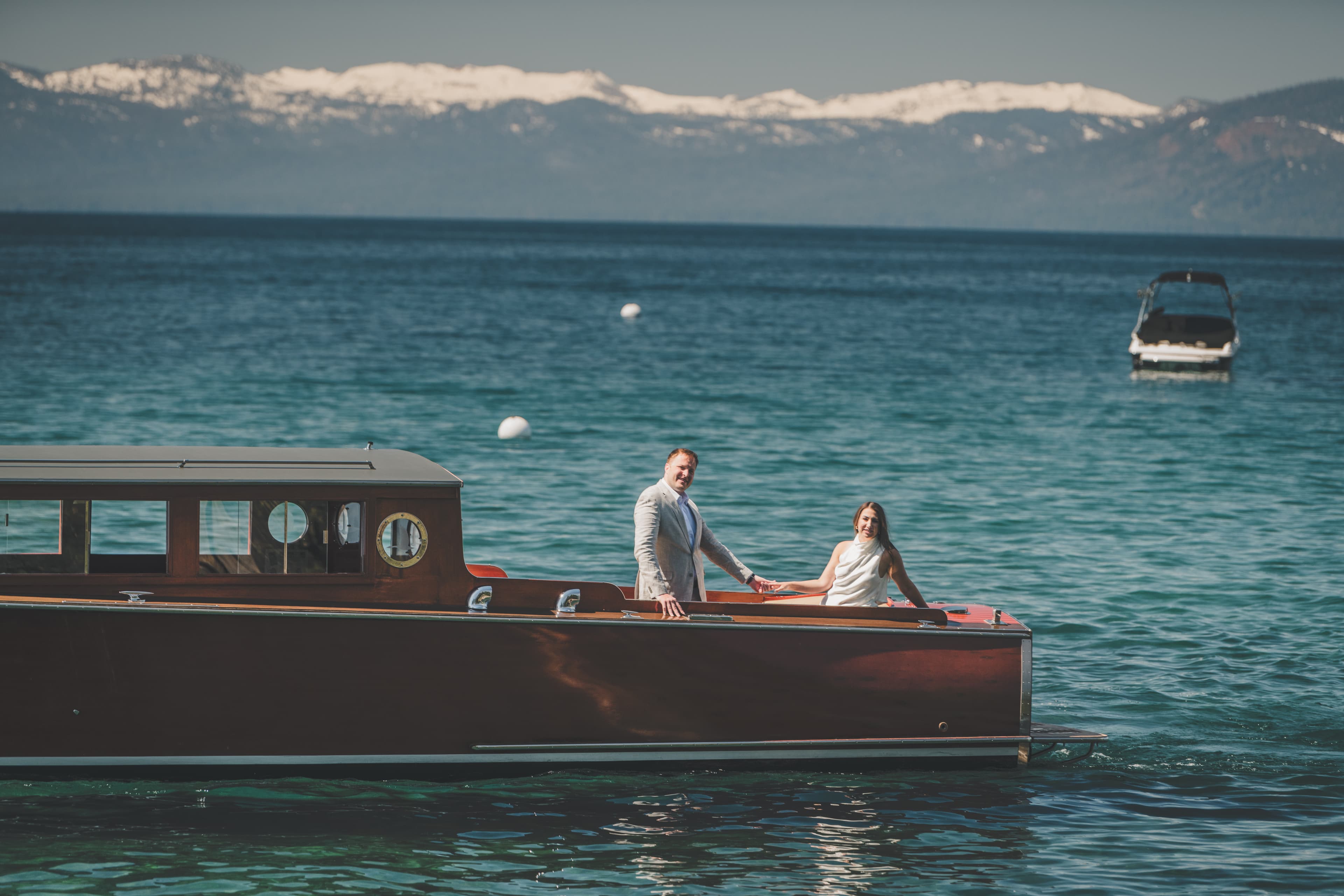 Hannah and Will standing on a classic mahogany boat with the snow-streaked Sierra Nevada across Lake Tahoe behind them
