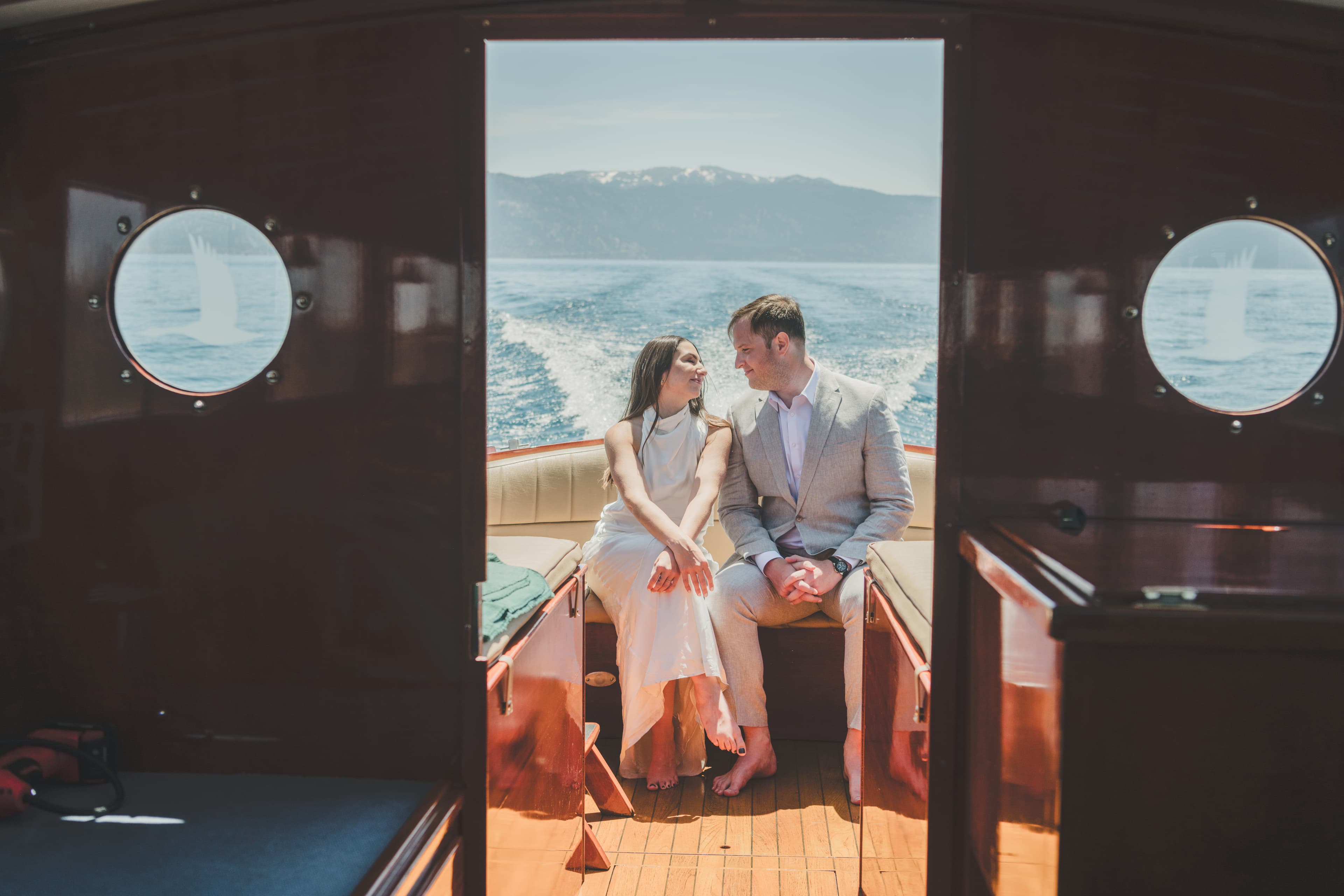 Hannah and Will framed by the cabin doorway of a wooden Tahoe boat, the lake stretching behind them through the portholes