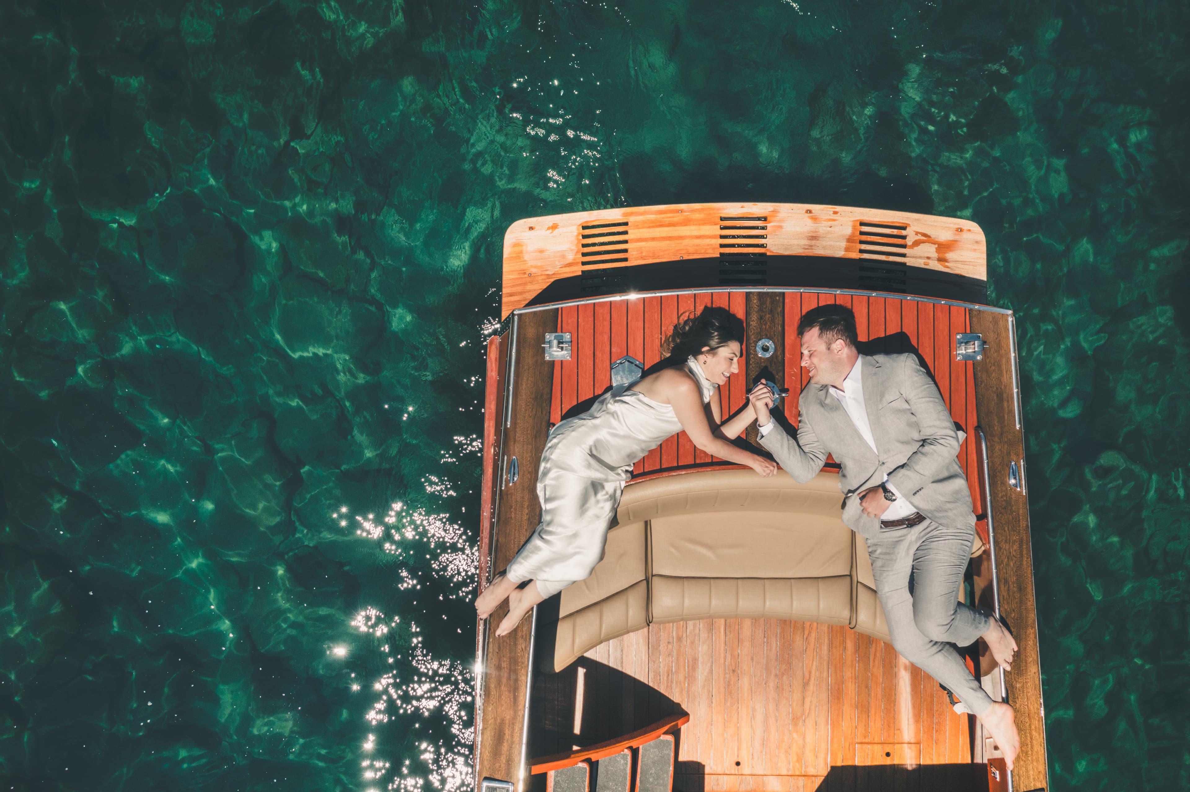 Aerial view down onto the wooden boat with Hannah and Will toasting at the stern over the green water of Lake Tahoe