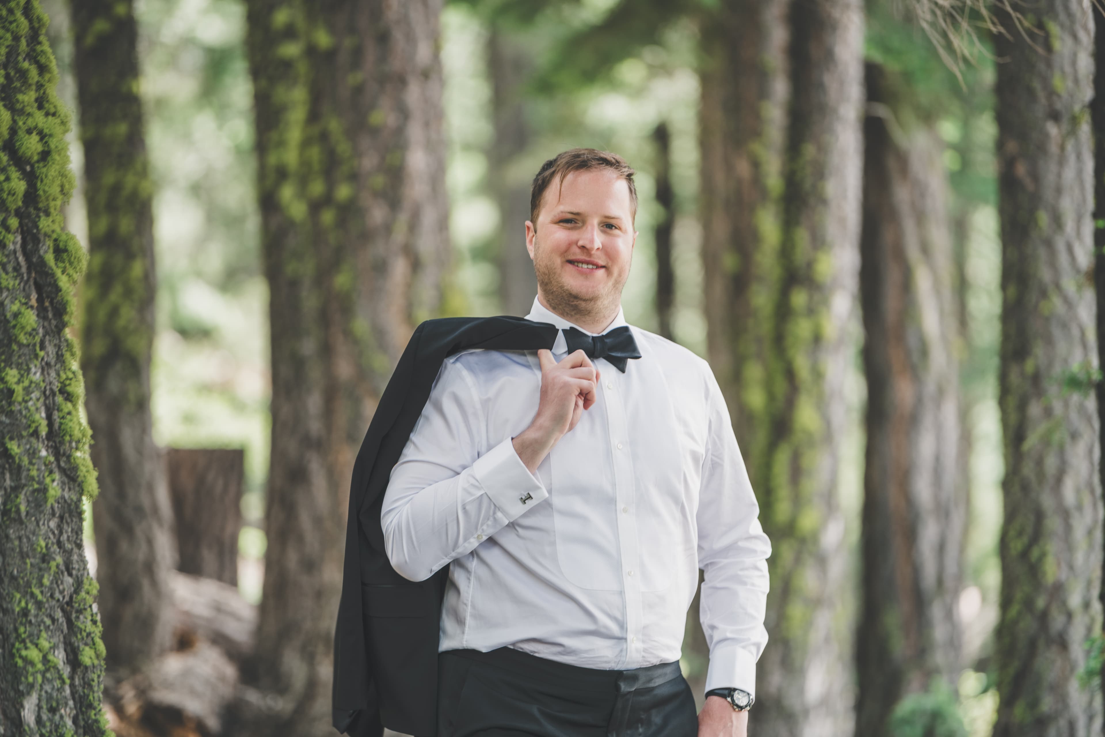 Will in white shirt and bow tie, jacket over his shoulder, in the tall pines above Everline Resort