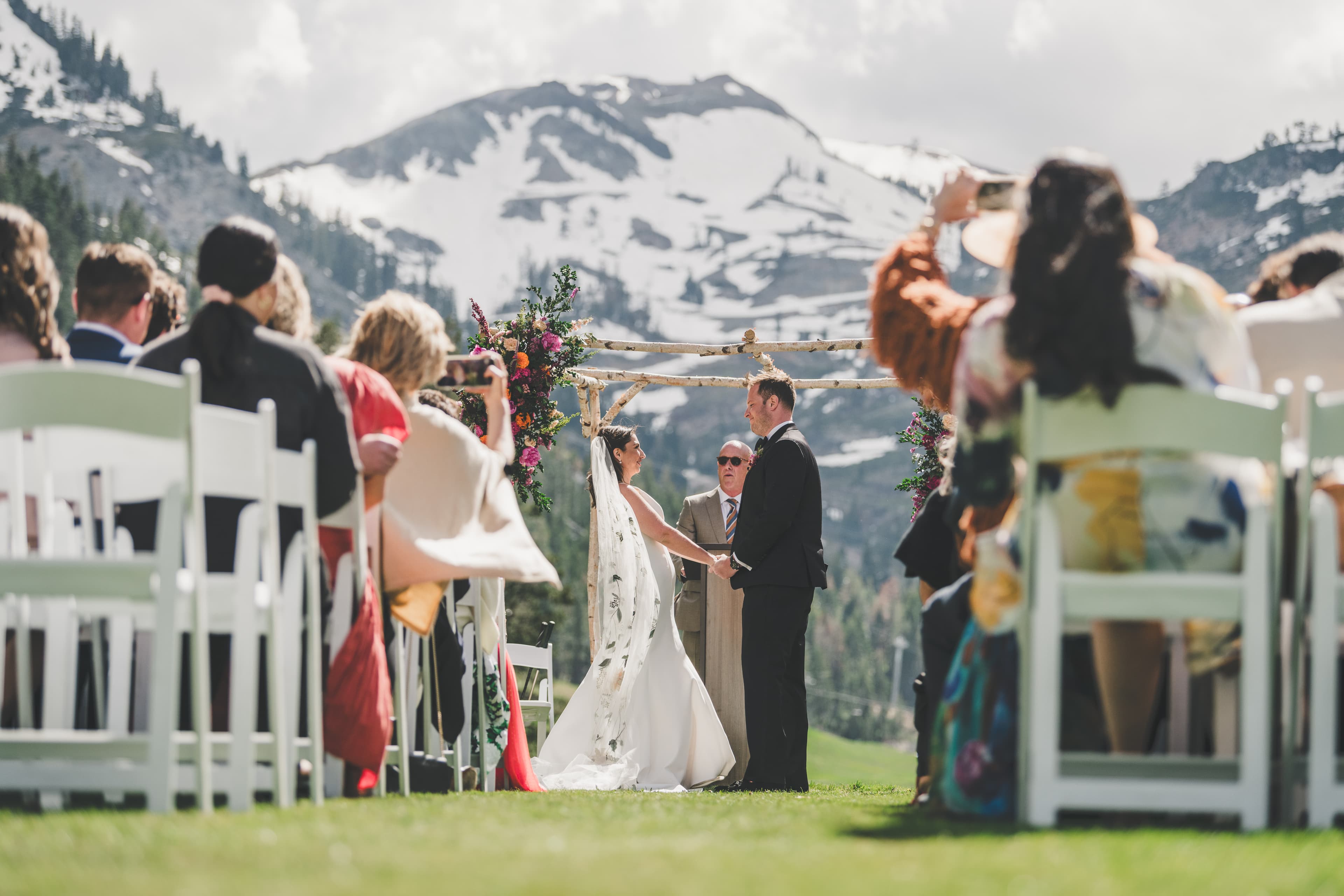 Hannah and Will at the altar mid-vows under the birch arbor with the snow-capped Sierra peaks behind them