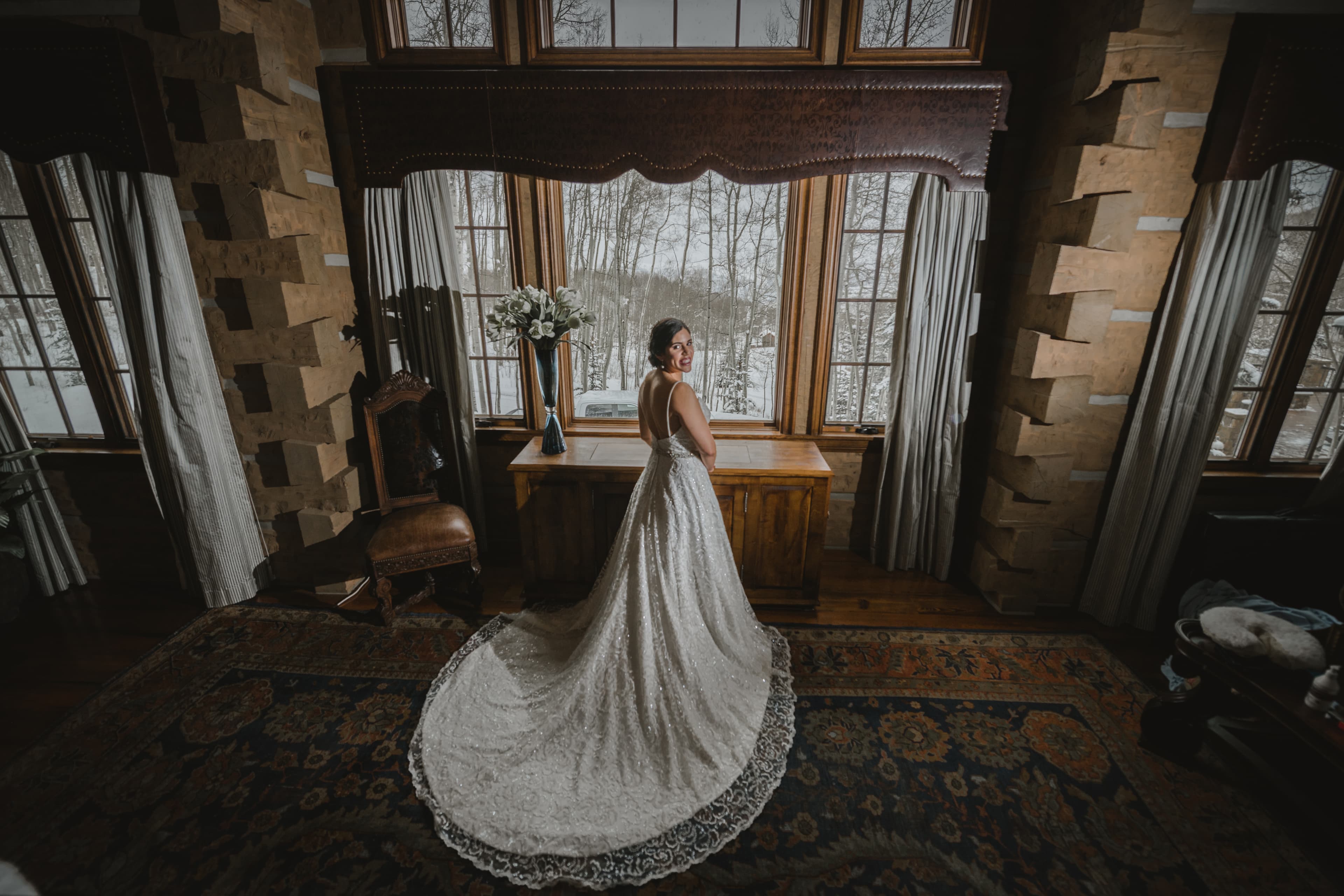 Sabrina in her wedding dress with snowy aspens visible through the window of the bridal suite