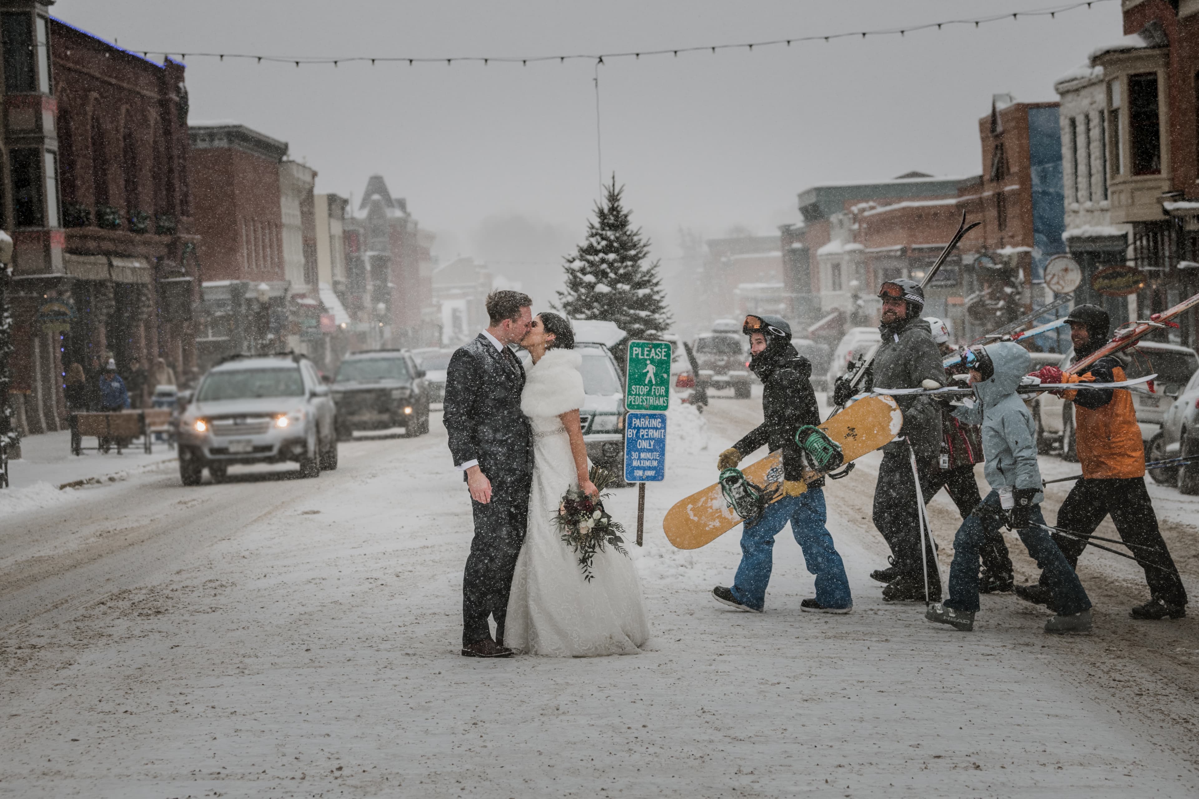 Sabrina and Patrick kiss on Telluride's main street as snowboarders pass by during a snowstorm