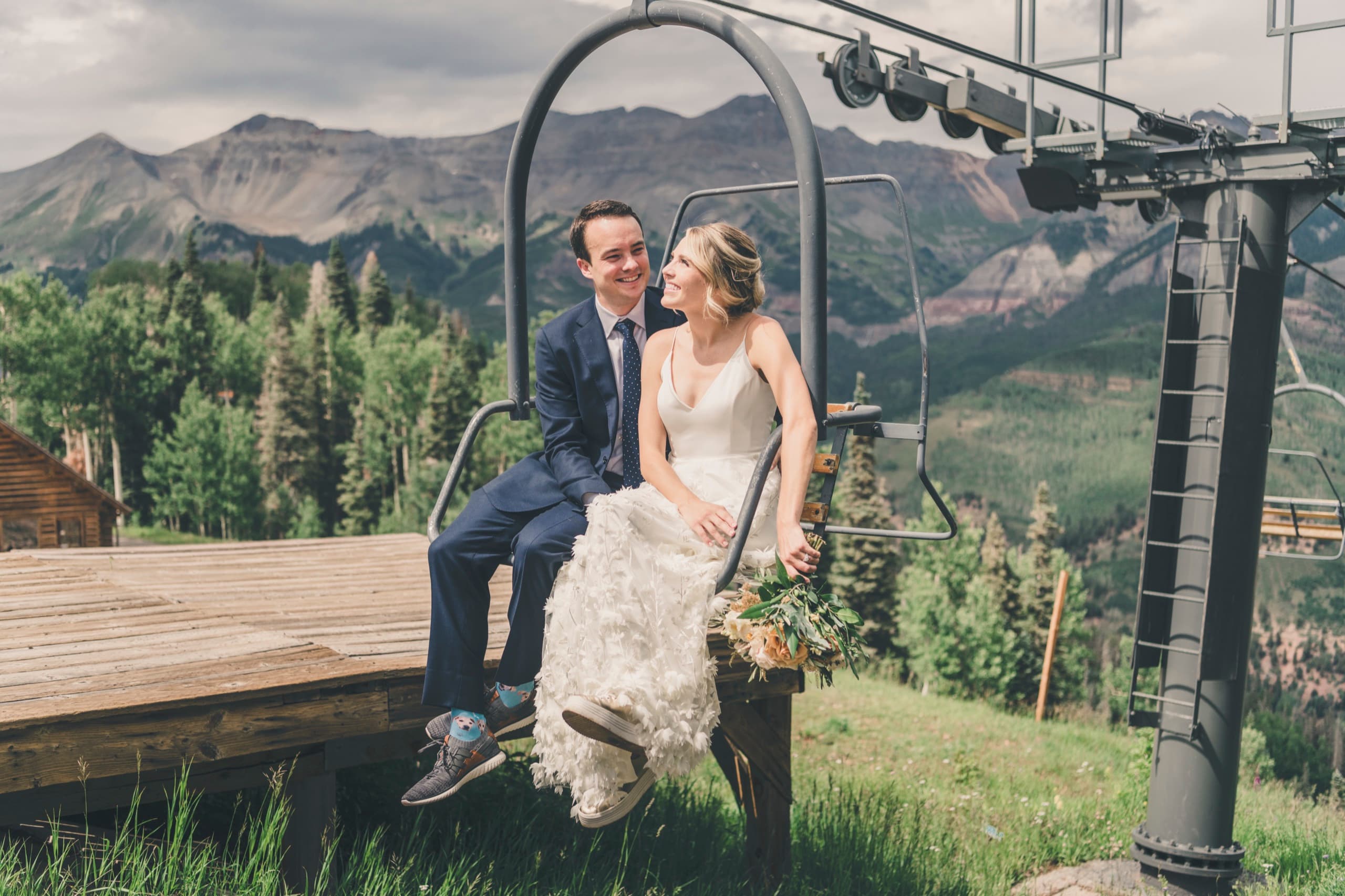 Meghan and Brent snuggled on the chairlift laughing at each other, mountain ridge and cabin behind