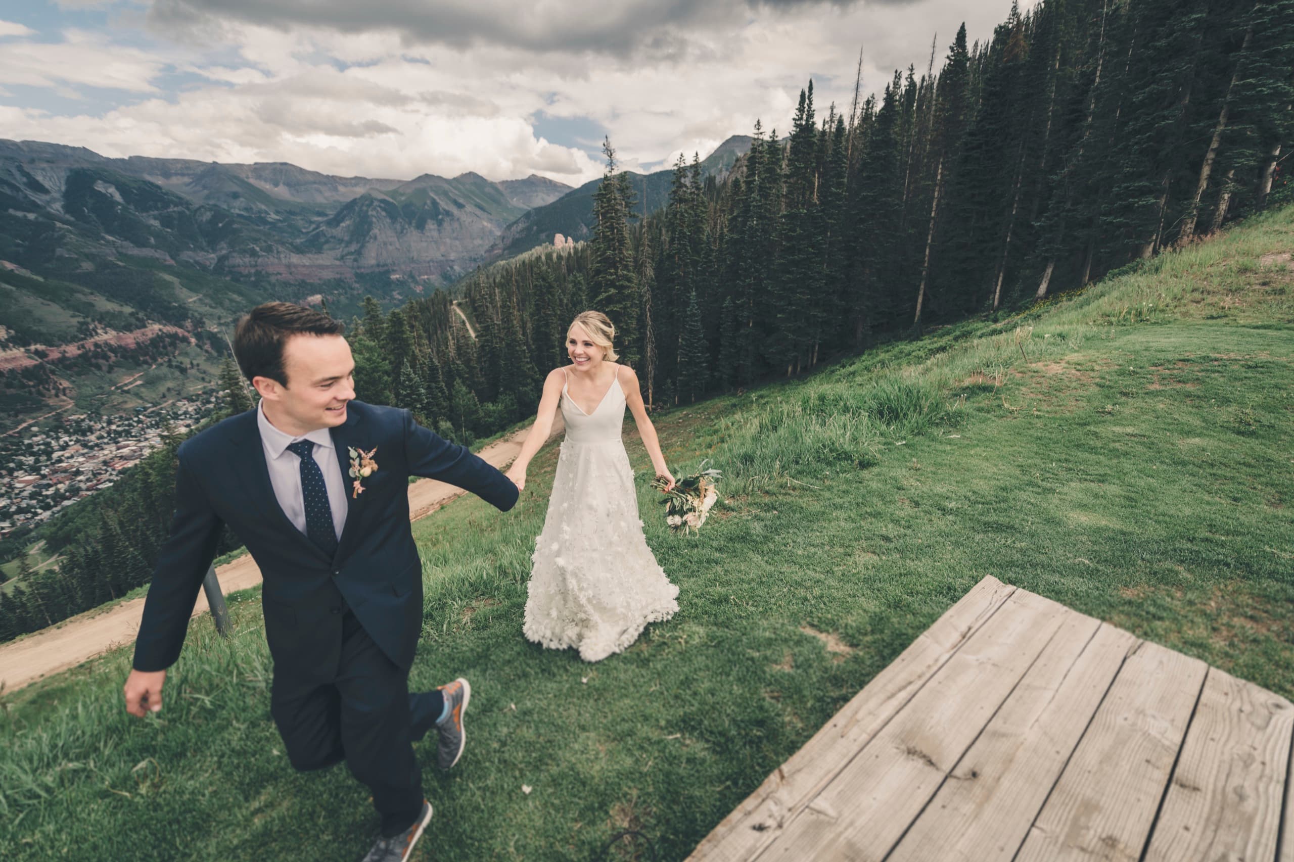 Meghan and Brent running hand in hand along the boardwalk on the Telluride ridge, dramatic clouds above