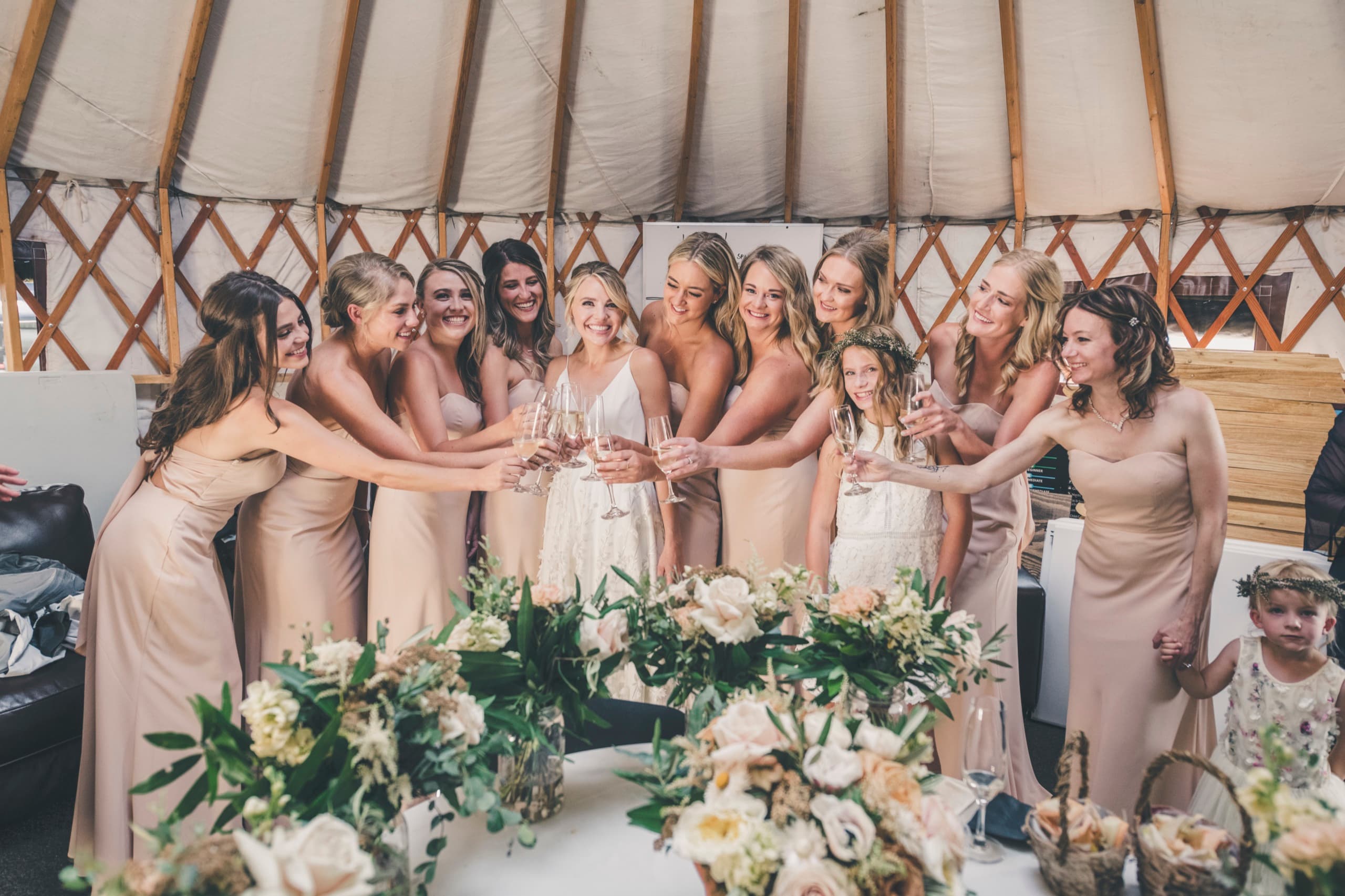 Meghan surrounded by bridesmaids in champagne dresses and the flower girl toasting in the prep yurt