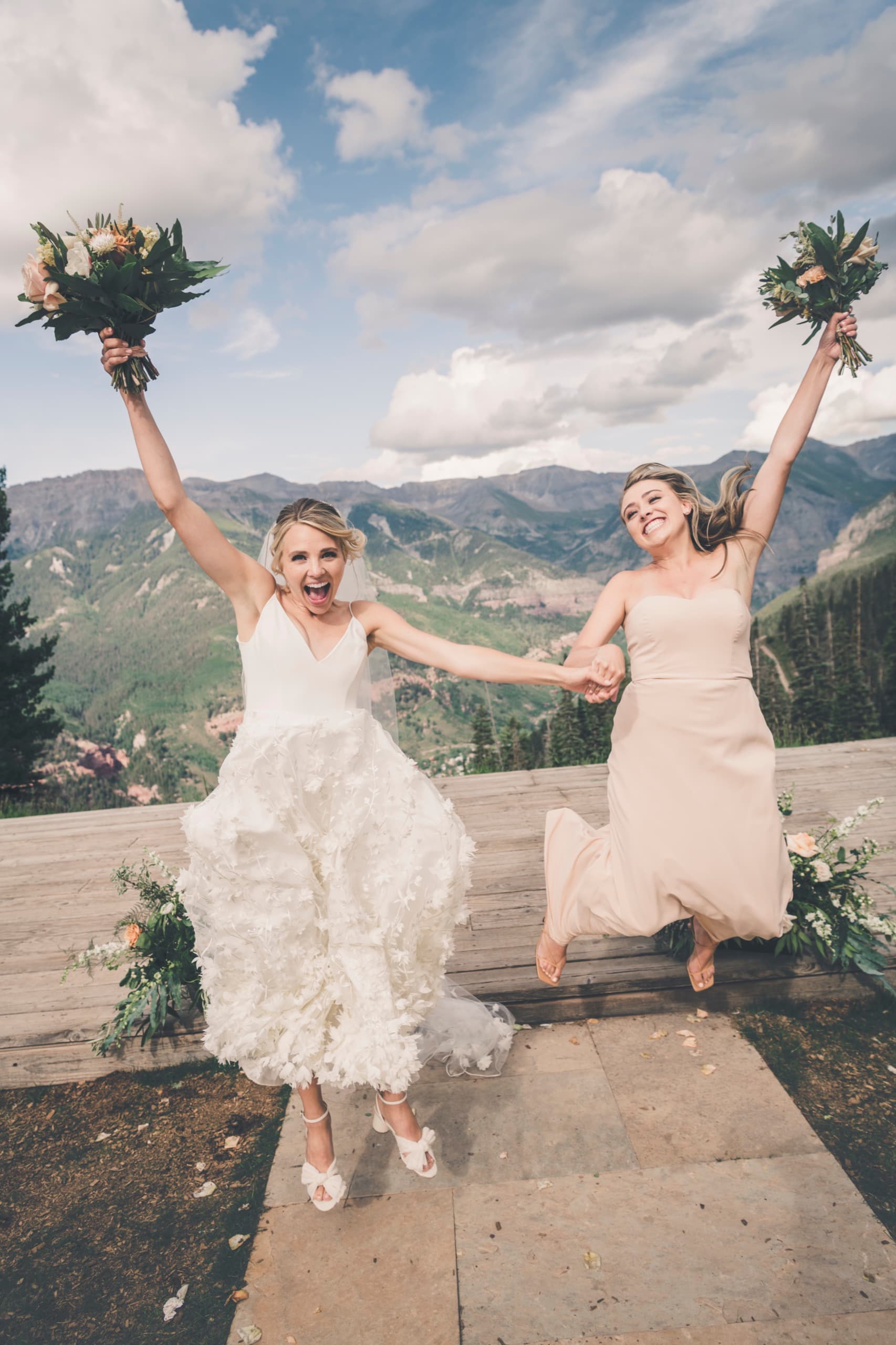 Meghan and a bridesmaid jumping mid-air with their bouquets raised on the Gorrono Ranch deck — pure storybook joy, no diva