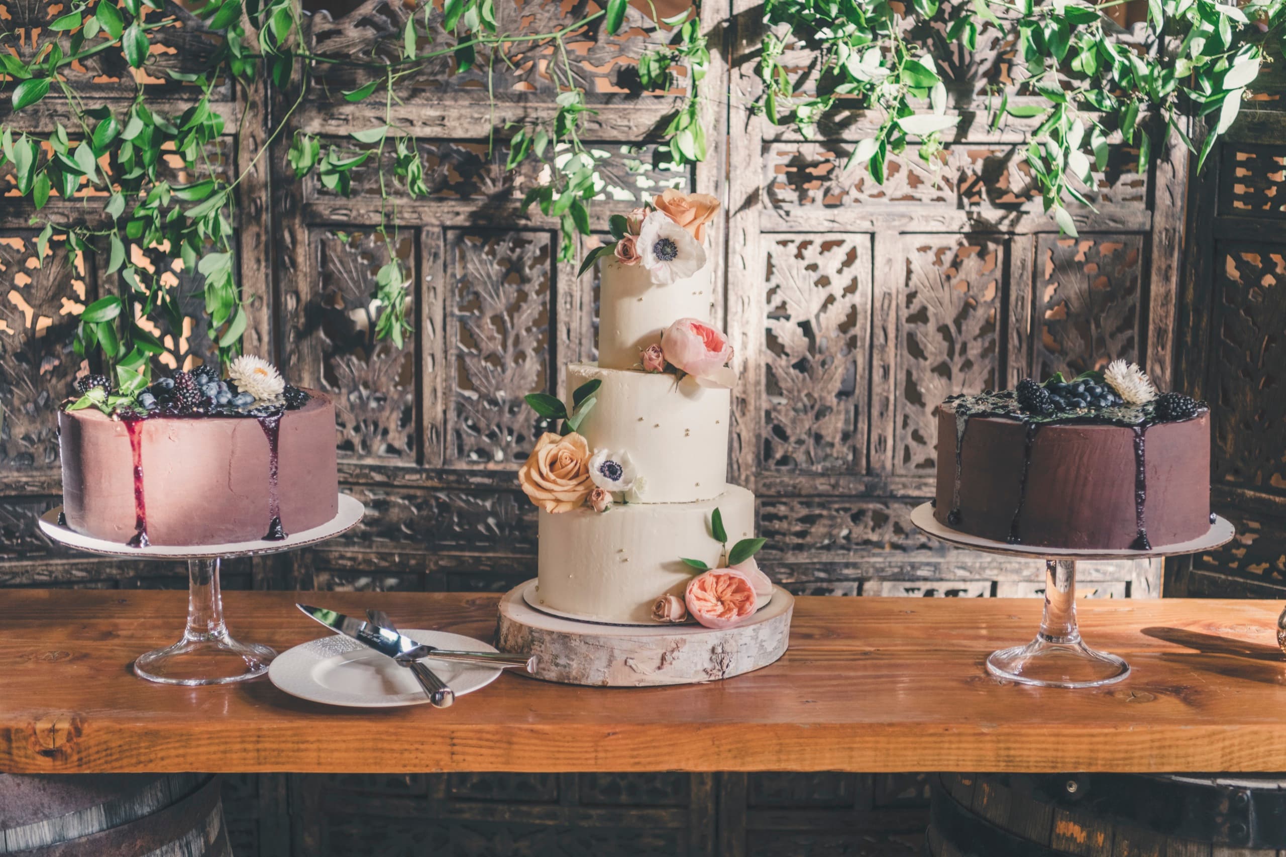 Three Meghan and Brent wedding cakes — purple, white, chocolate — on a wood plank against rustic carved wood