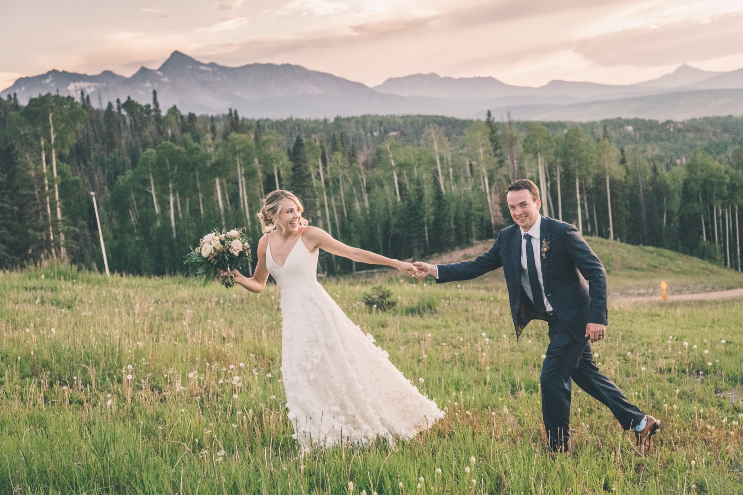 Meghan and Brent walking hand in hand through a Telluride wildflower meadow at sunset, San Sophia ridge behind them