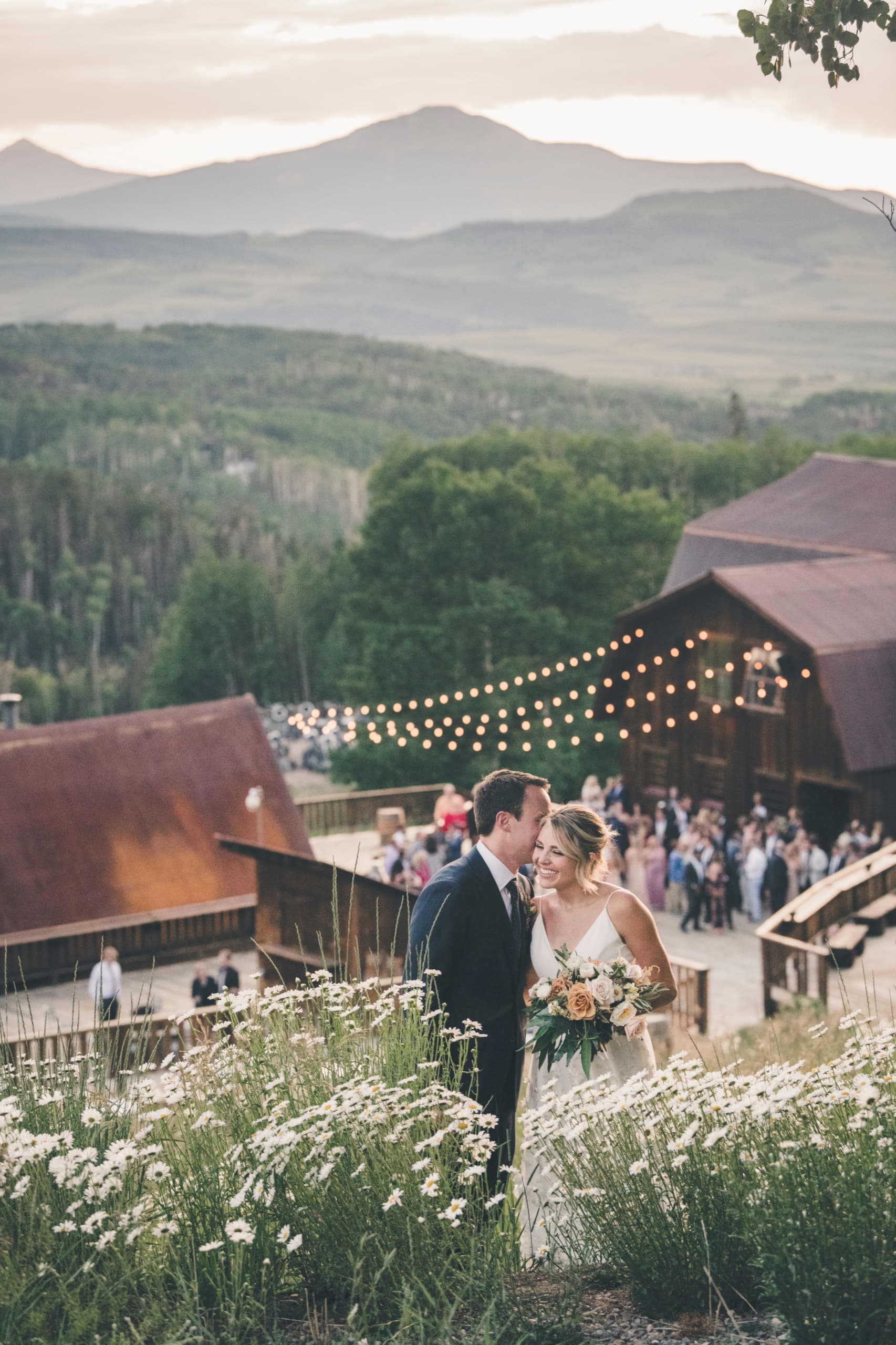 Meghan and Brent in a daisy meadow with the lit Gorrono lodge and string lights behind them at golden hour