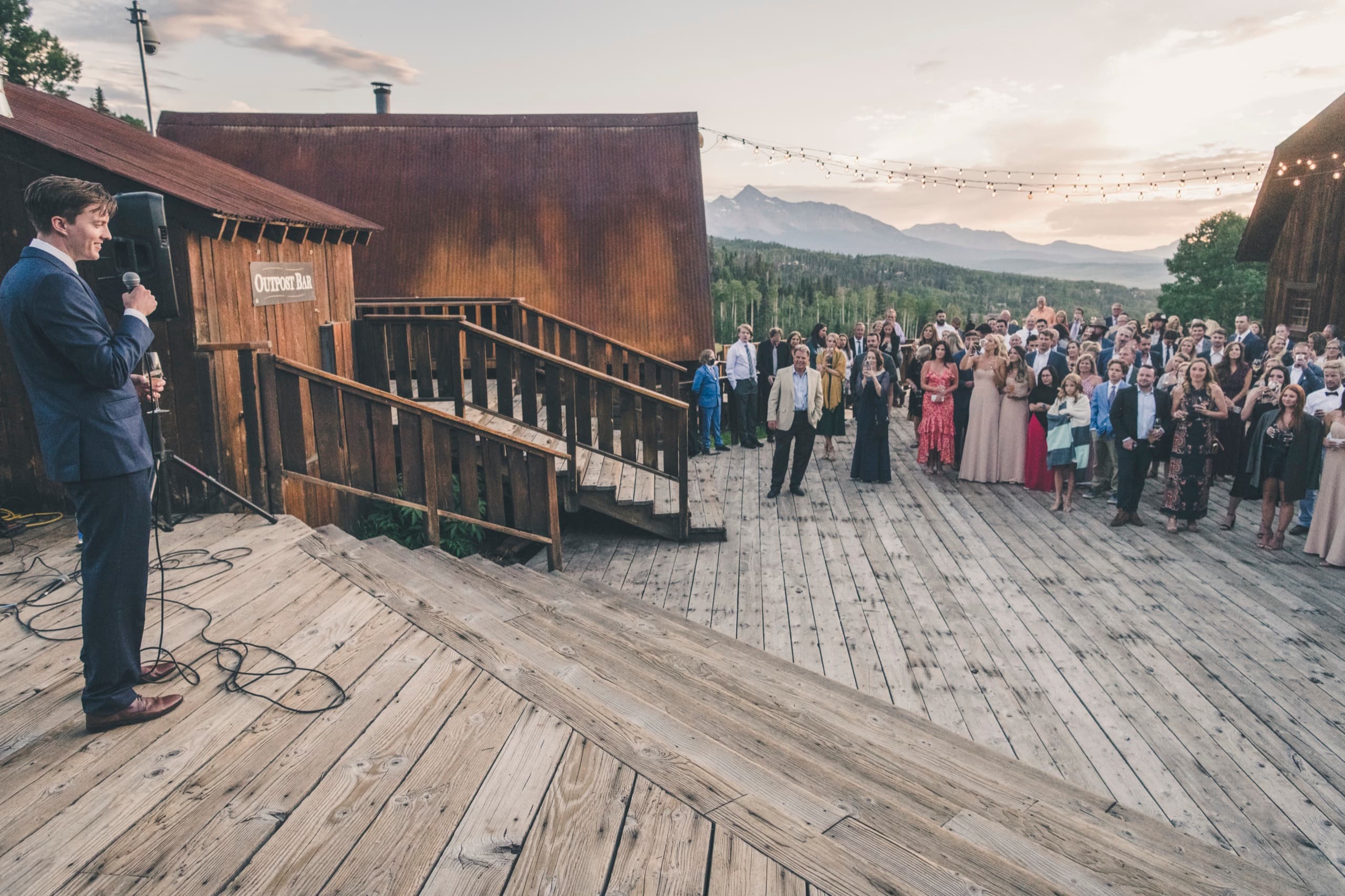 Wide of the Gorrono Ranch deck reception — speaker giving a toast to guests under string lights at golden hour