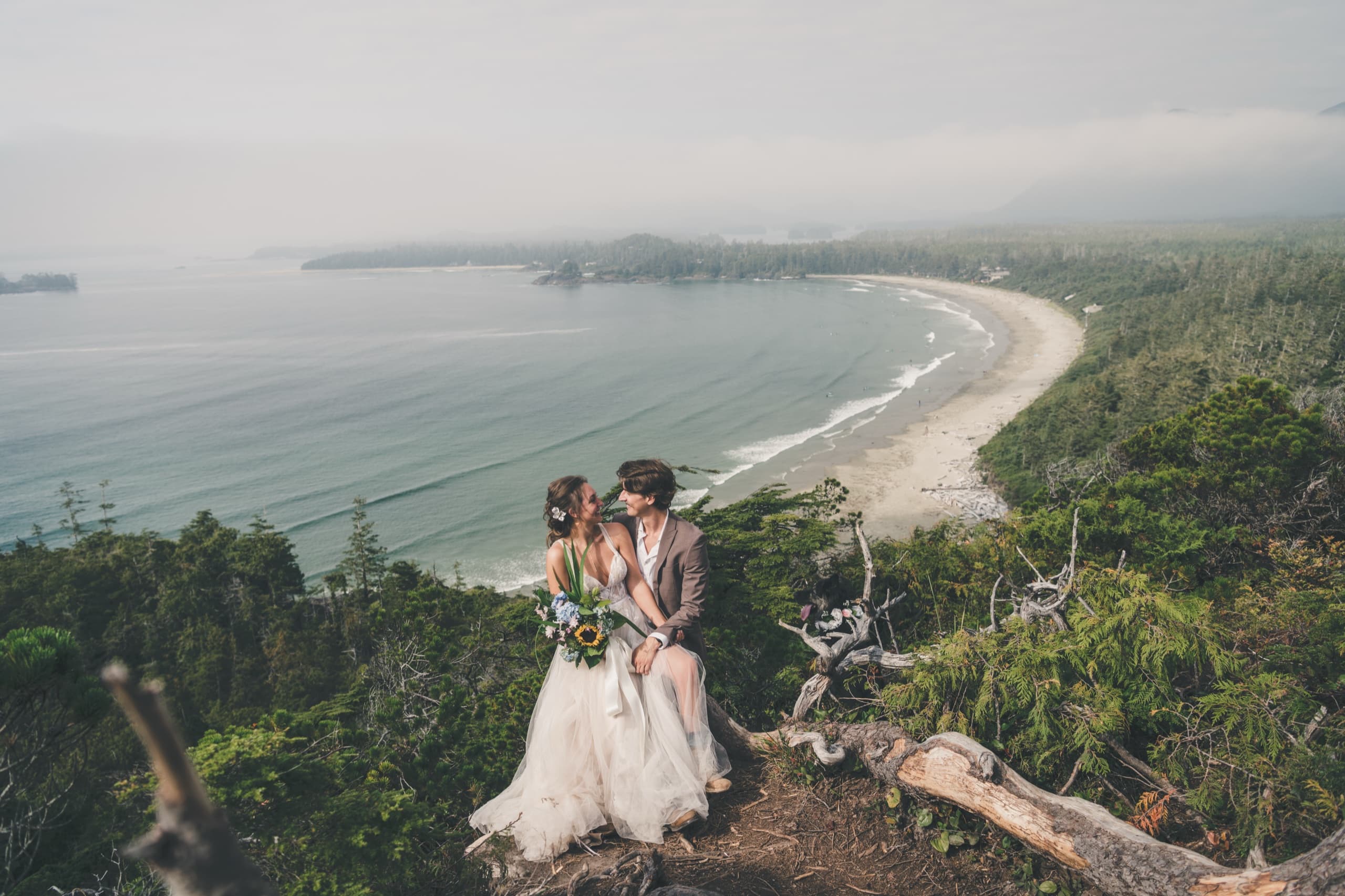 Rachel and Peter seated on a cliff overlooking the foggy Tofino bay and crescent beach