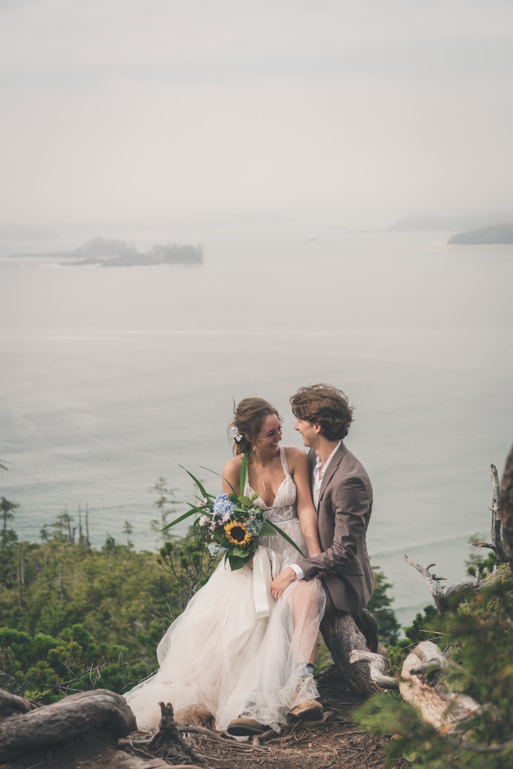 Rachel laughing on a driftwood cliff edge with Peter, the Pacific and offshore islands behind