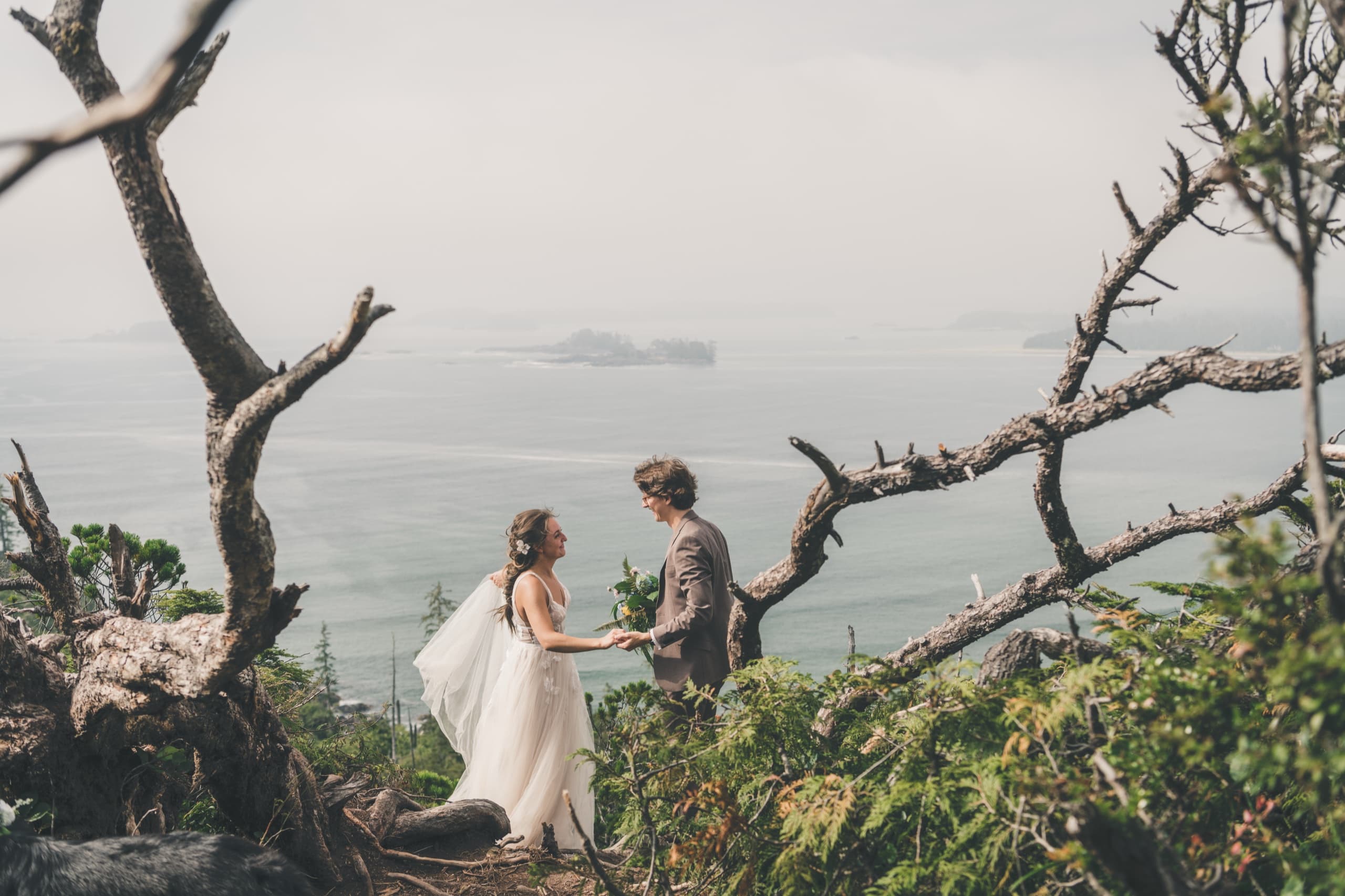 Rachel and Peter holding hands on a windy cliff framed by gnarled driftwood branches above the Pacific