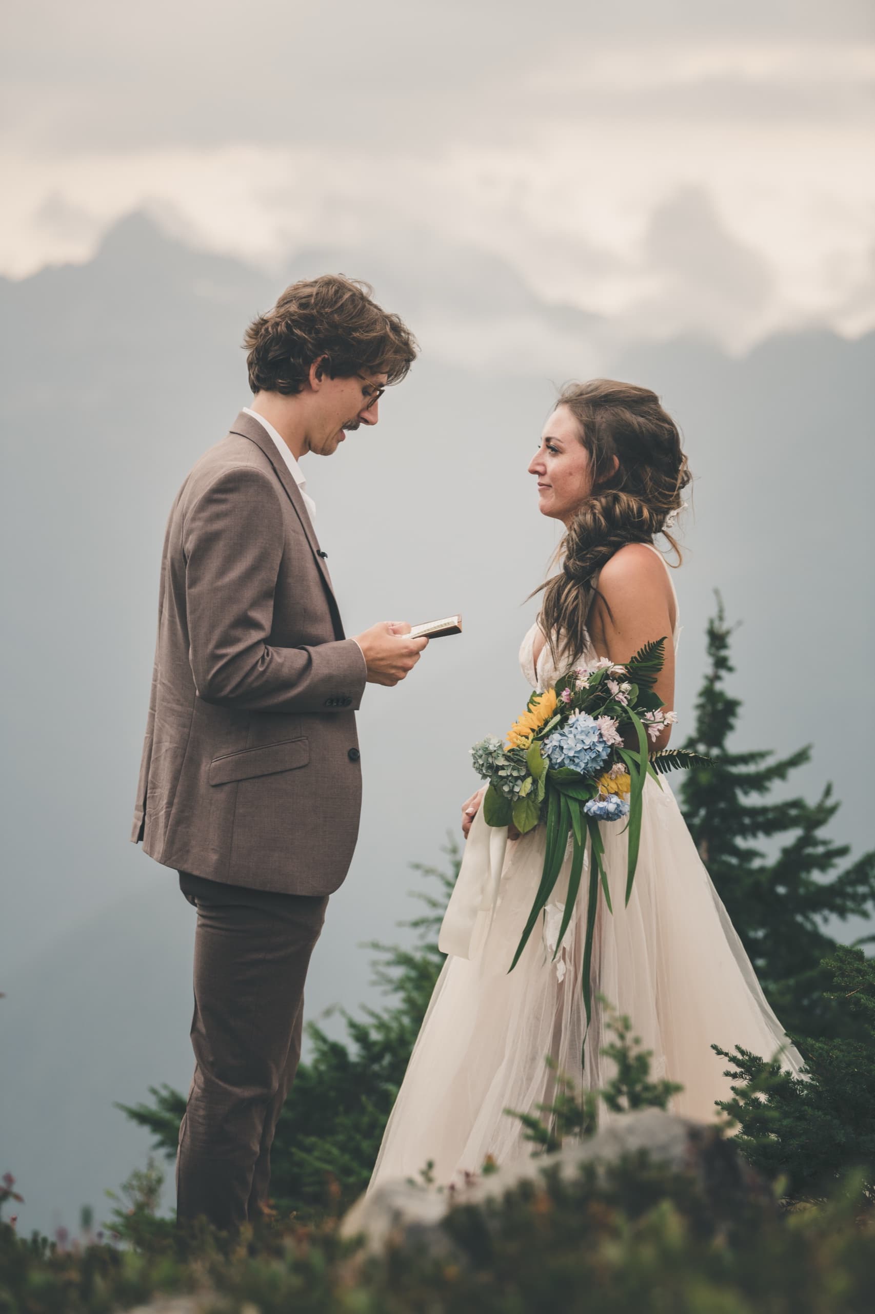 Rachel and Peter face to face reading their vows on an alpine peak with cloud-shrouded peaks behind