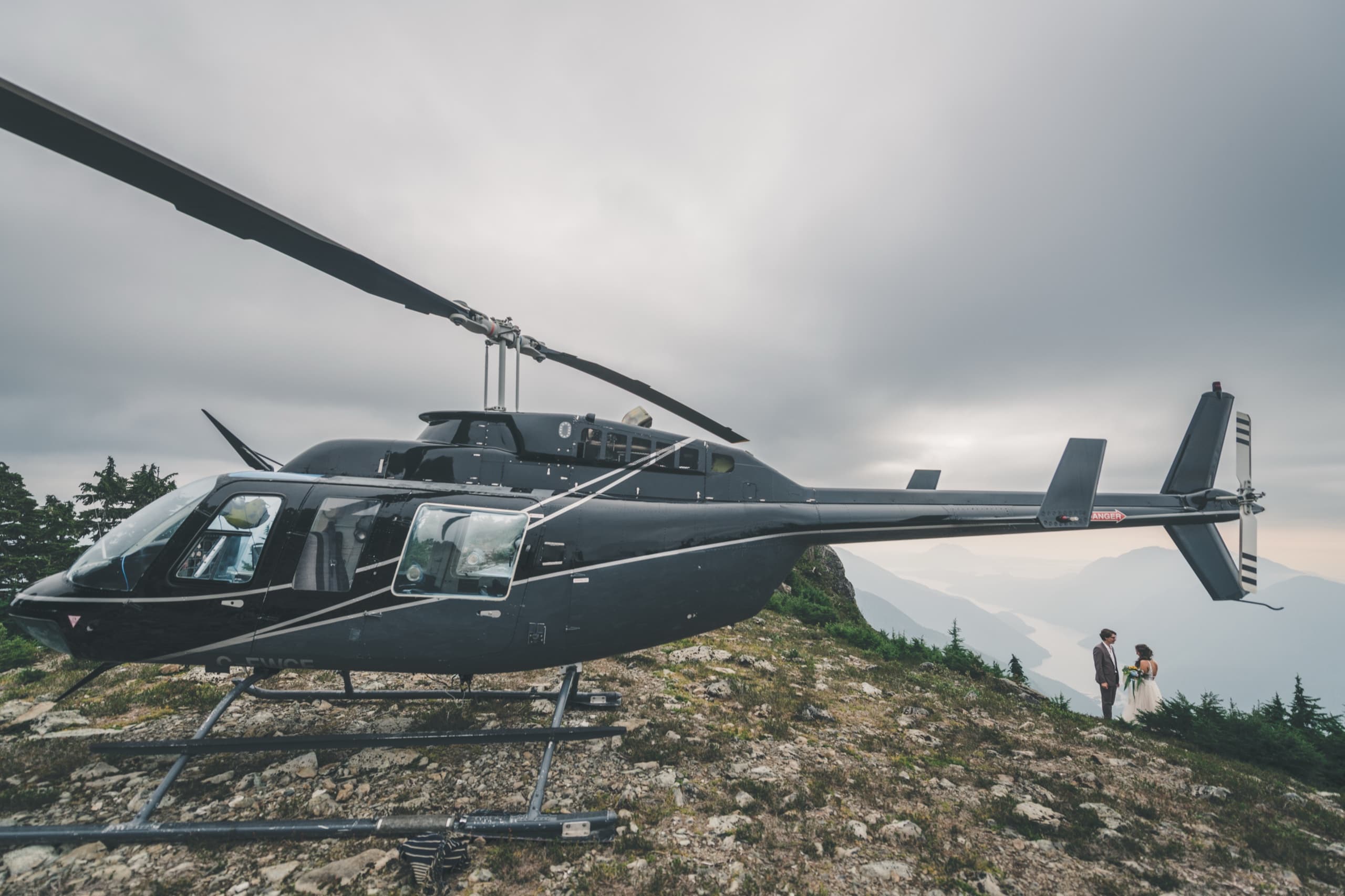 The chartered black helicopter parked on a rocky alpine ridge, Rachel and Peter tiny beside it