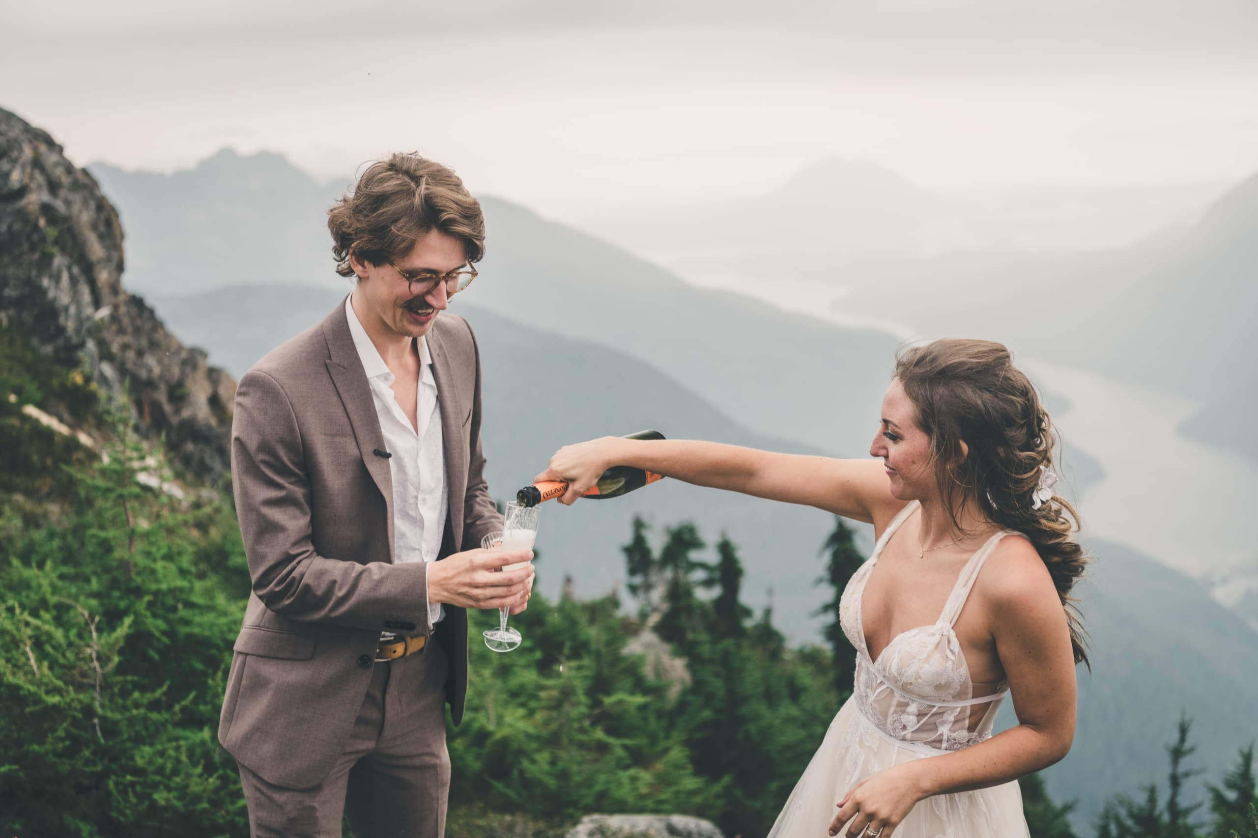 Rachel pouring champagne into Peter's flute on a mountaintop, the fjord behind them