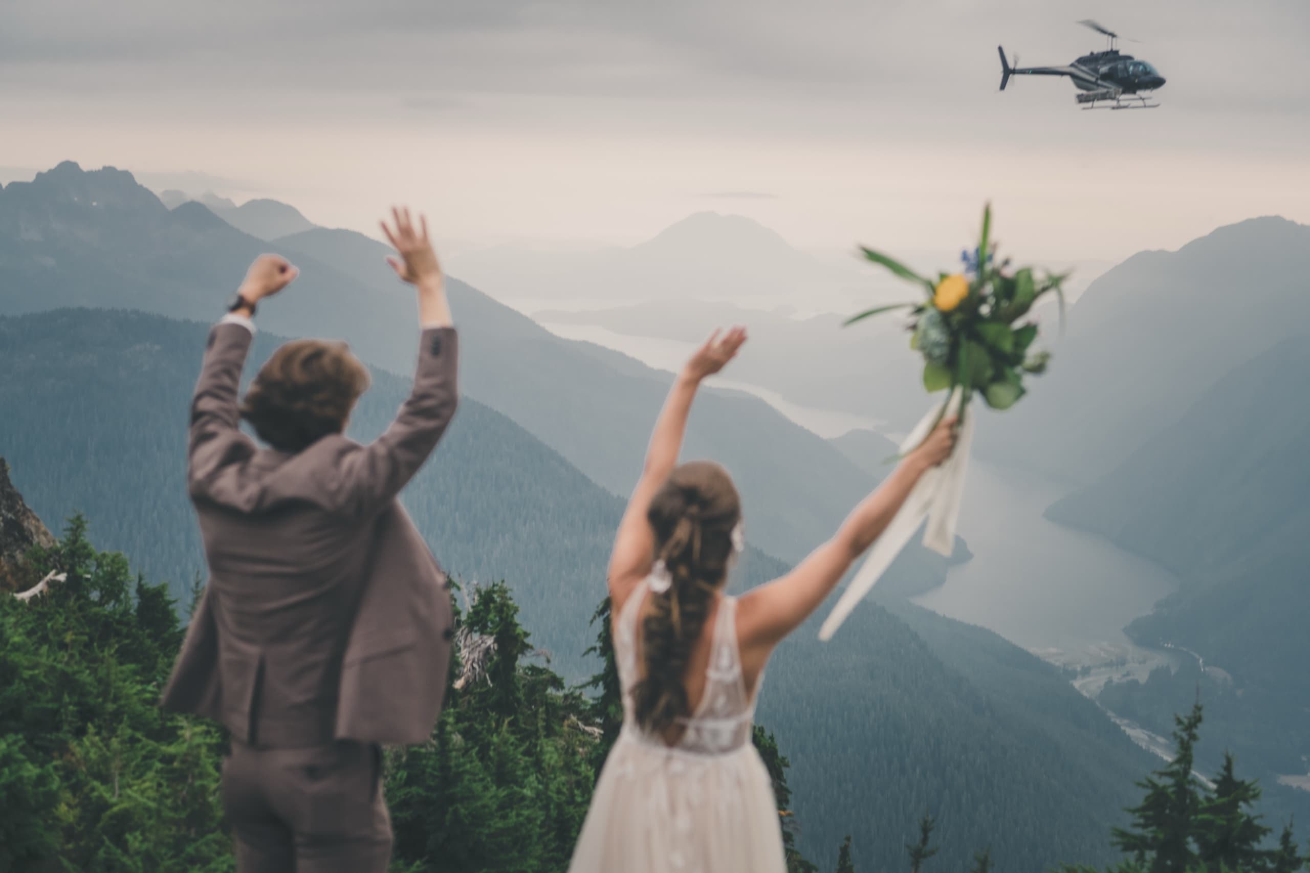 Rachel and Peter cheer with arms raised as the helicopter lifts off into the mountains behind them