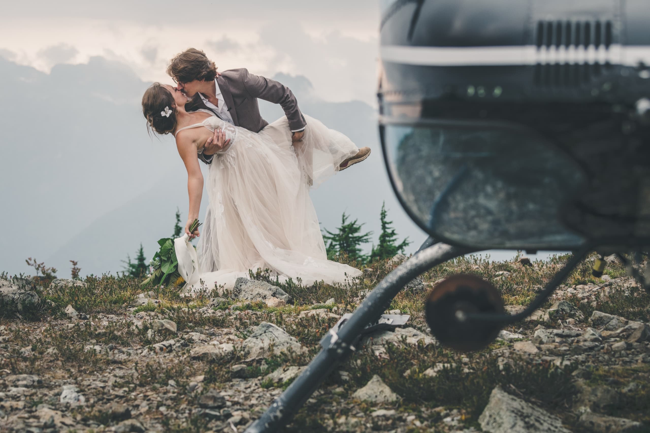 Rachel and Peter dip kiss with the helicopter nose in the foreground of the alpine ridge