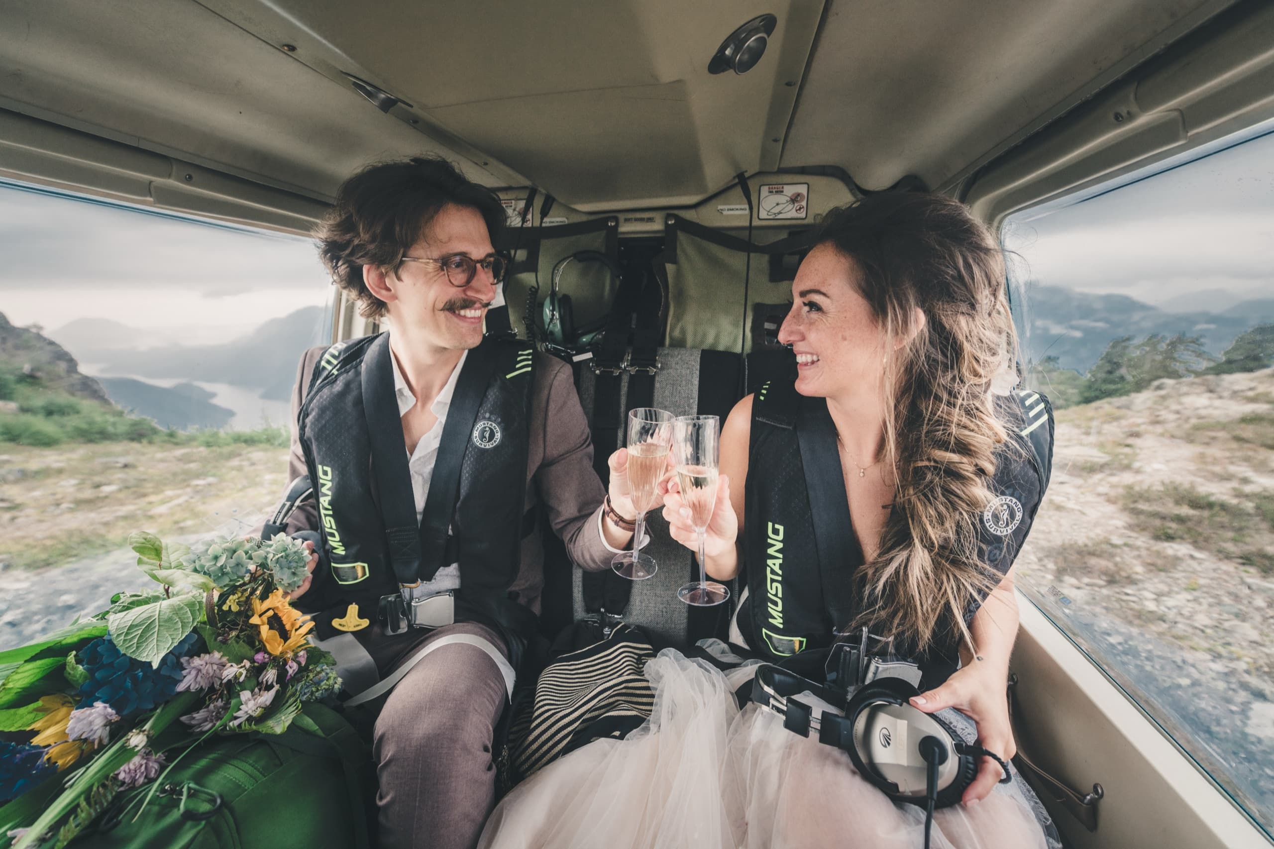 Rachel and Peter inside the helicopter cabin in life vests, toasting champagne with mountains through the window