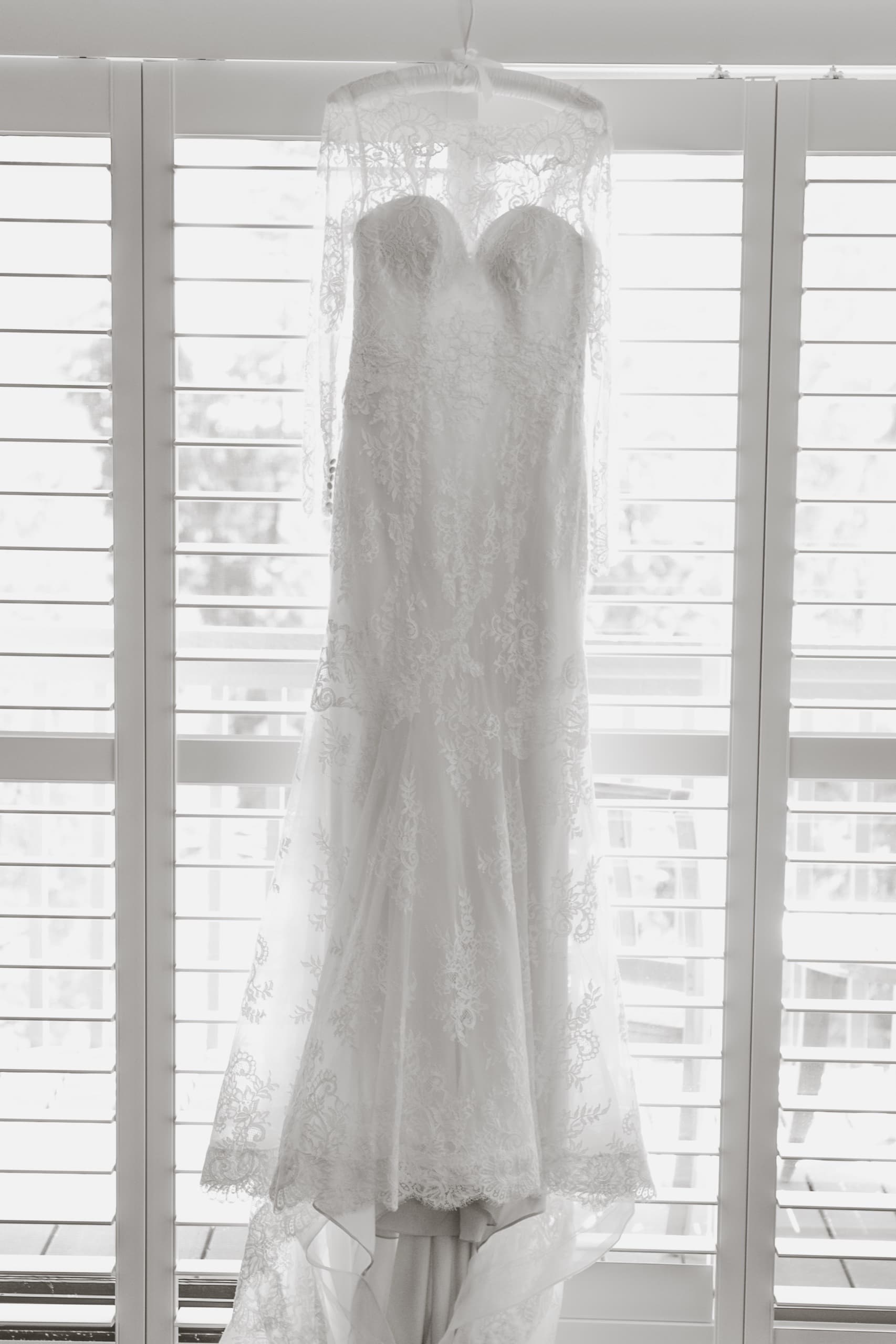 Jolly's lace wedding dress hanging in the window with shutters in soft natural backlight, black and white