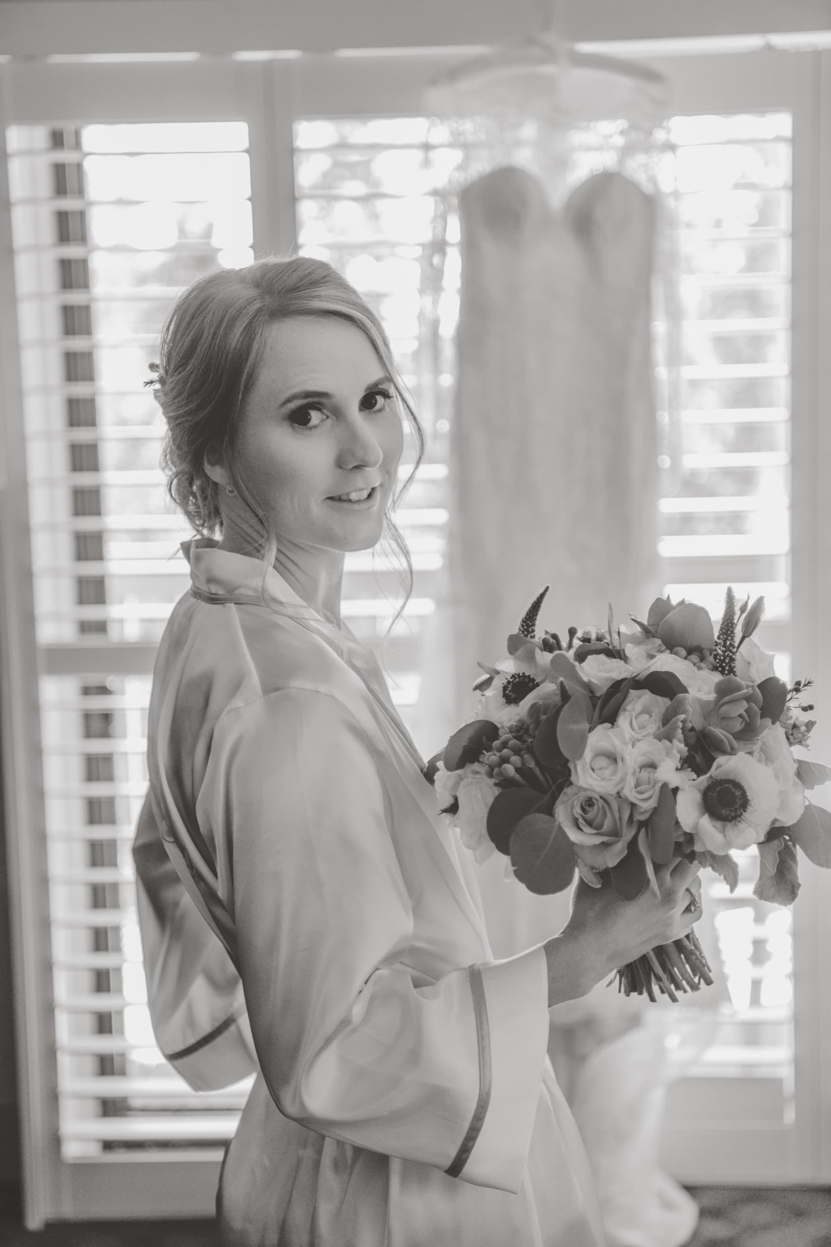 Jolly in her robe holding her bouquet with her dress hanging behind her, getting-ready window light