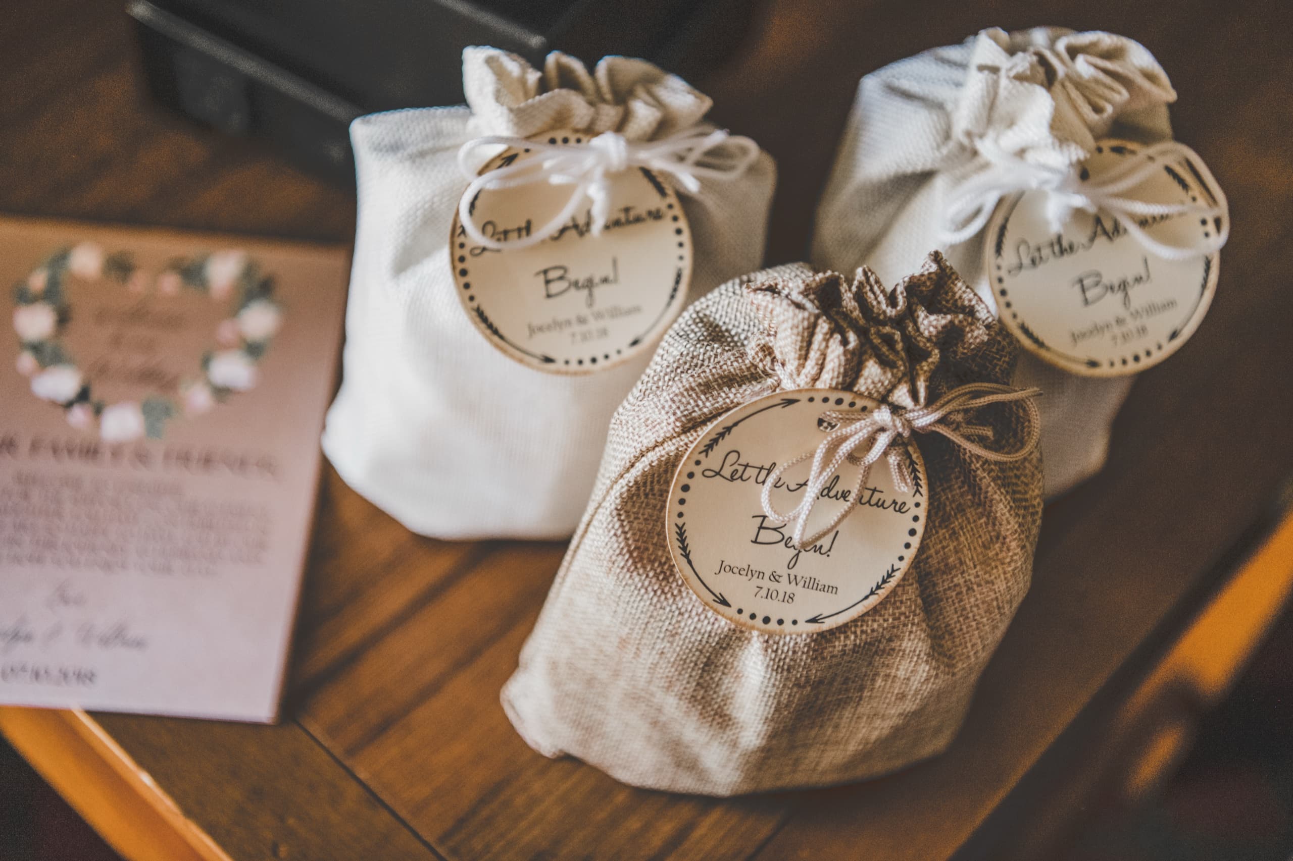 Three burlap Little Adventure Begins favor bags with the wedding invitation in warm indoor light