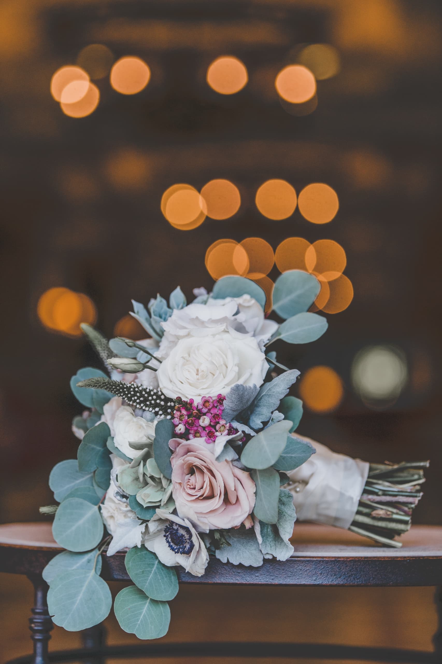 Jolly's bridal bouquet of roses, eucalyptus, and anemone on a chair with warm bokeh string lights behind