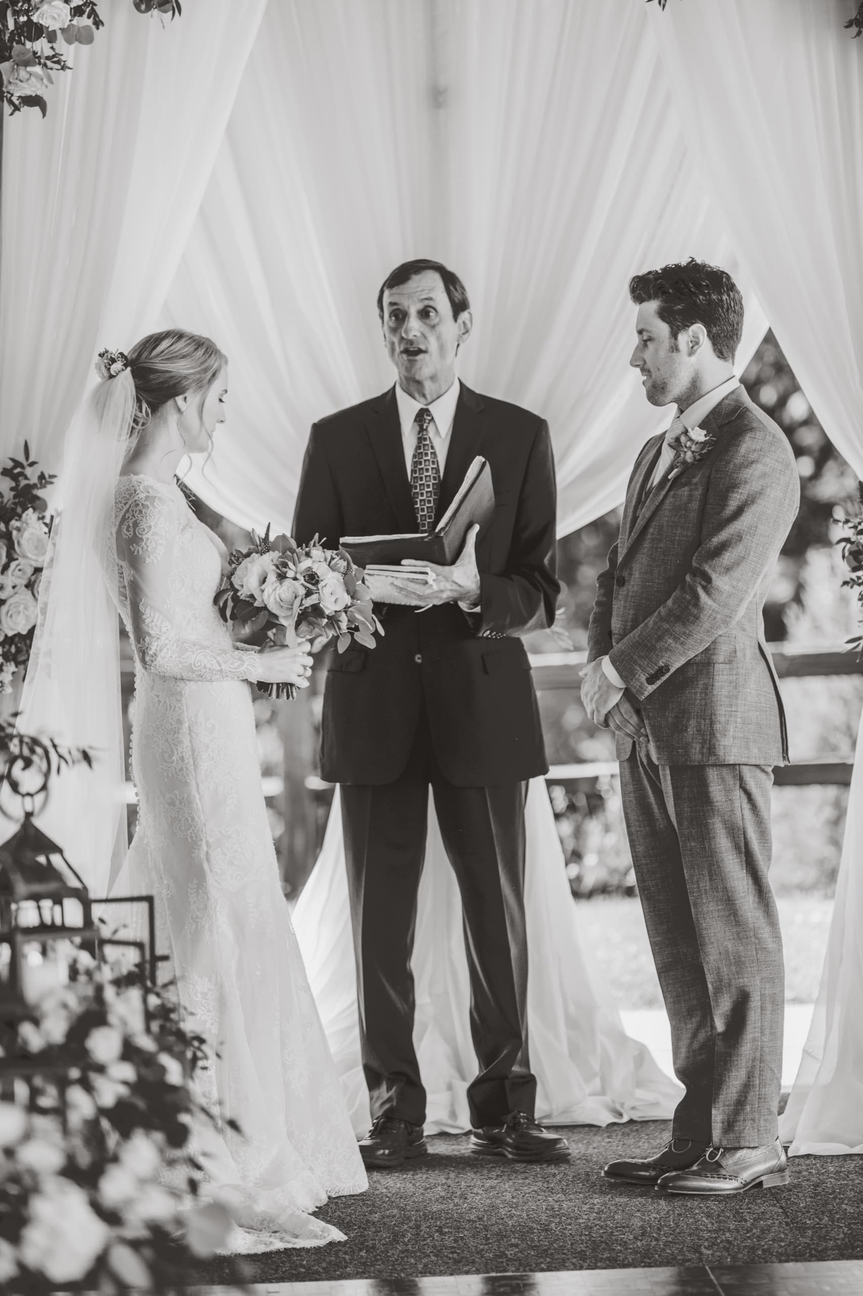 Jolly and Billy face their officiant under the white drapes of the ceremony pavilion in black and white