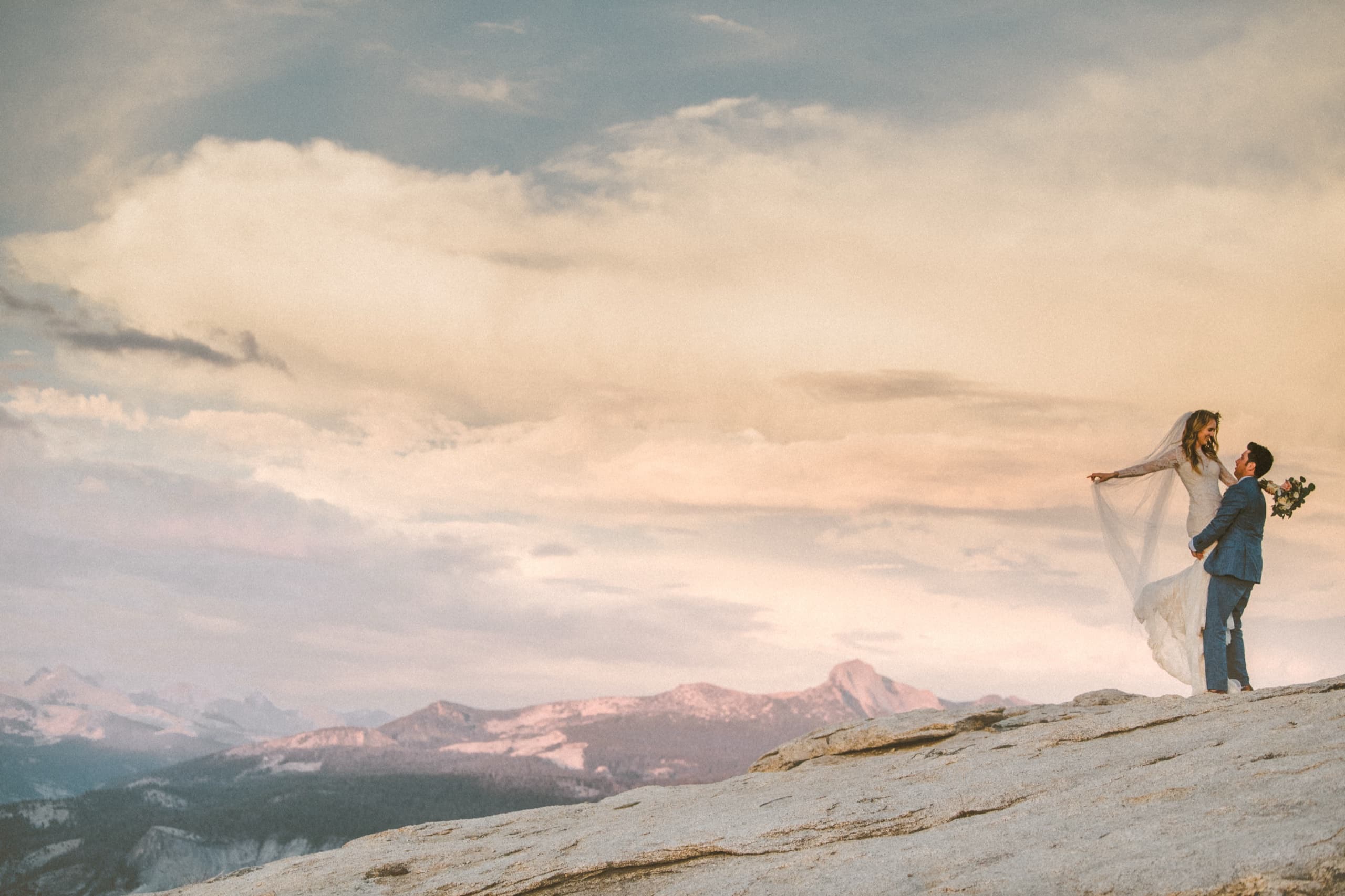 Wide shot of Billy lifting Jolly into the air on the granite dome with the Sierra peaks and pastel sky behind
