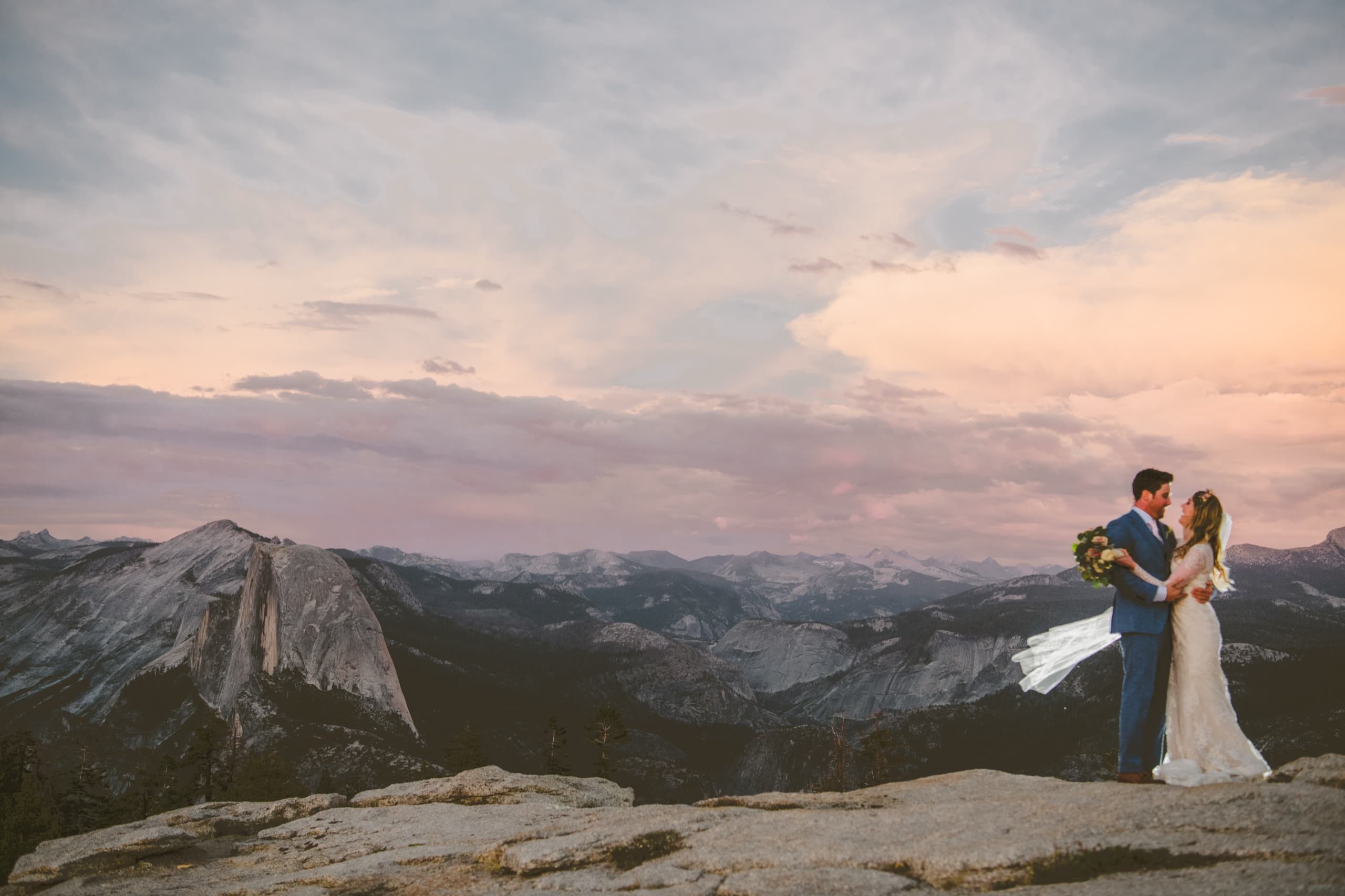 Jolly and Billy embracing on Sentinel Dome with Half Dome and a pink-sky Sierra panorama behind them